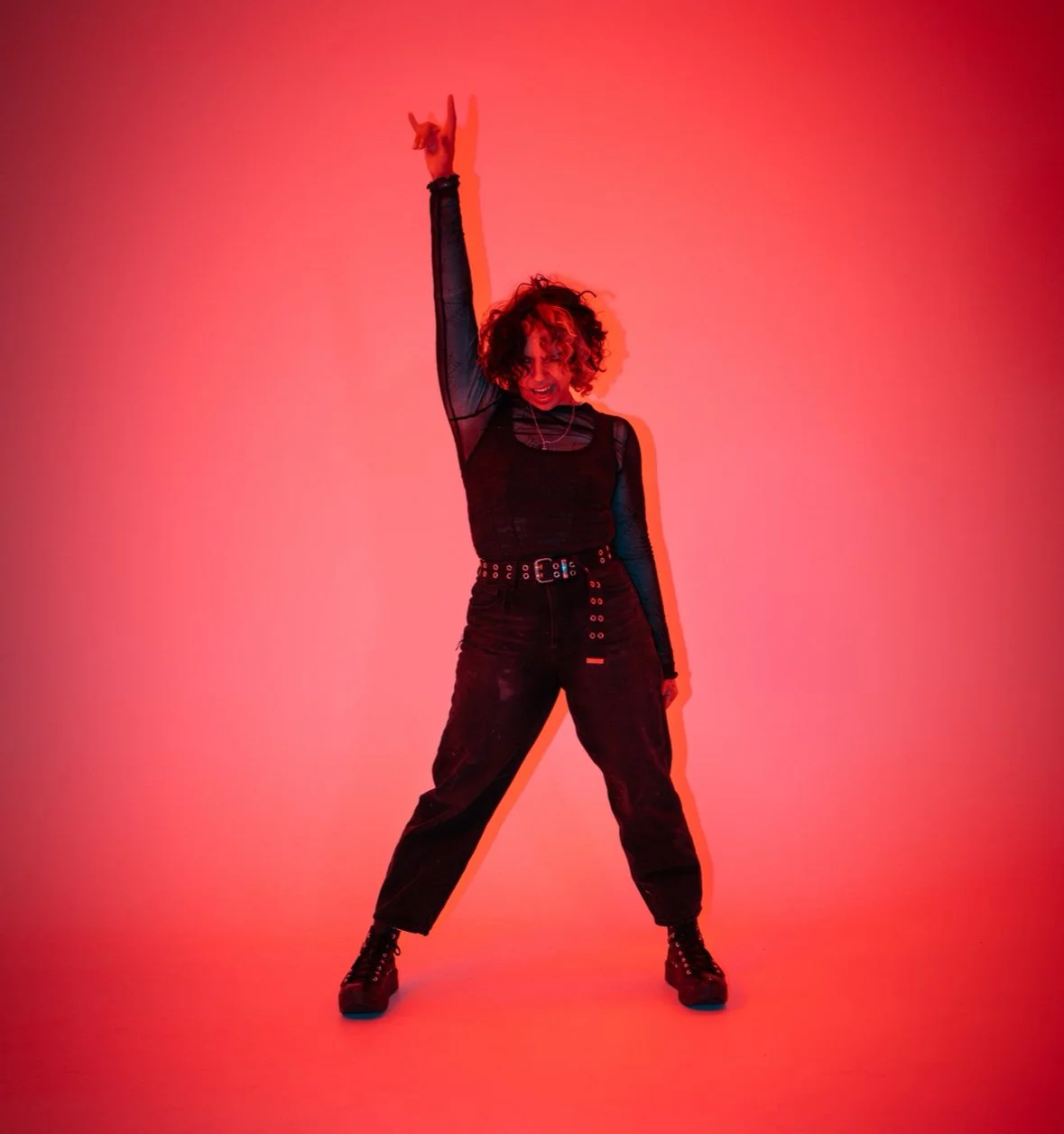 A woman with curly hair wearing all black, standing against a red gradient background, striking a confident pose with one arm raised and making a rock 'n' roll hand gesture.