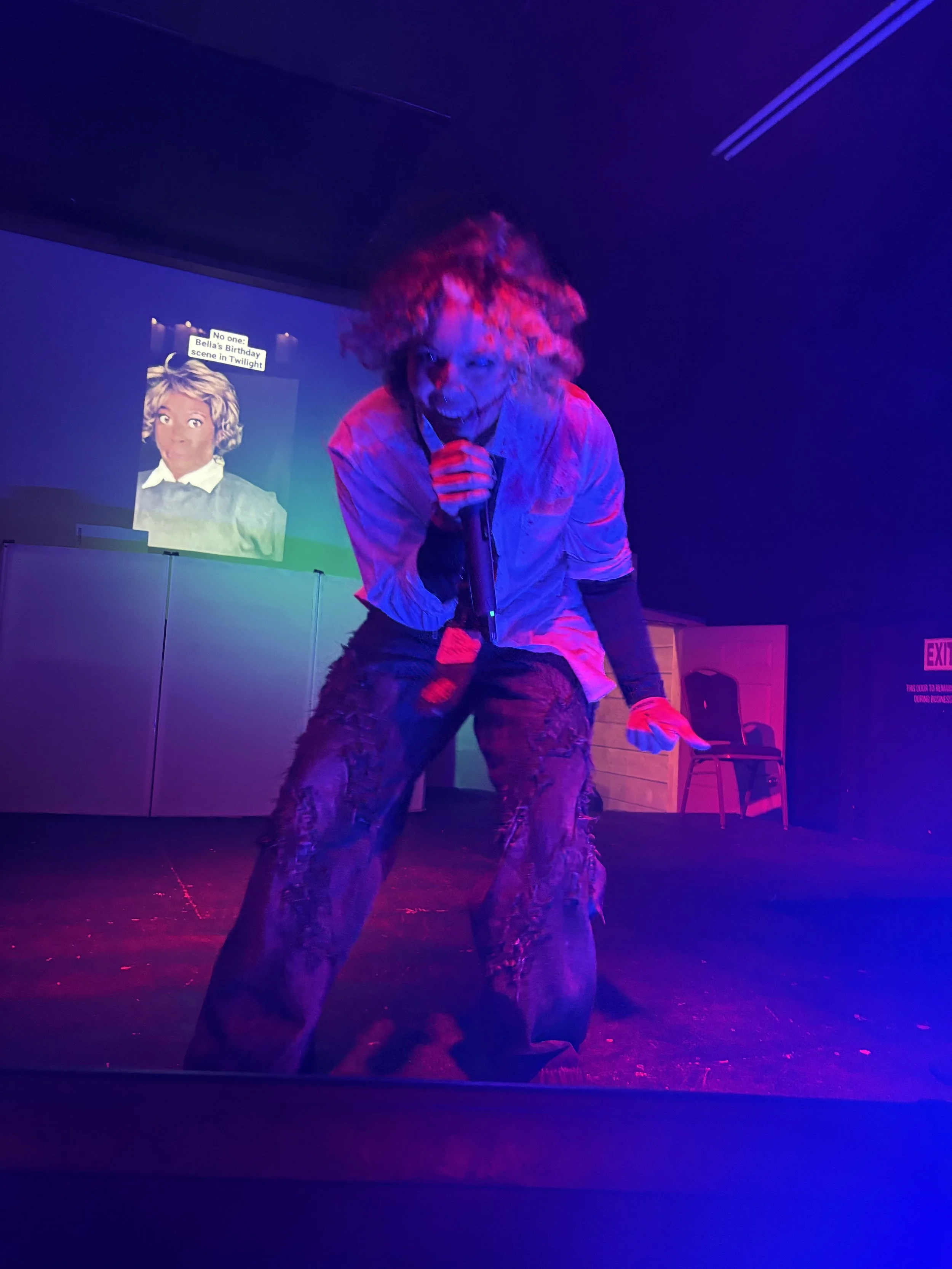 Person performing on stage with messy hair, holding a microphone, in front of a colorful light and screen projection showing a woman with curly hair.