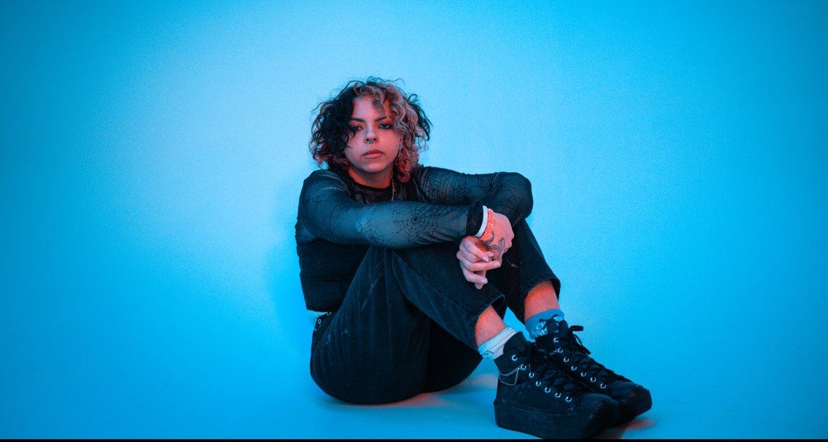 Young woman sitting on the floor against a blue background, with curly hair, dressed in black clothing with lace sleeves, wearing black high-top sneakers.