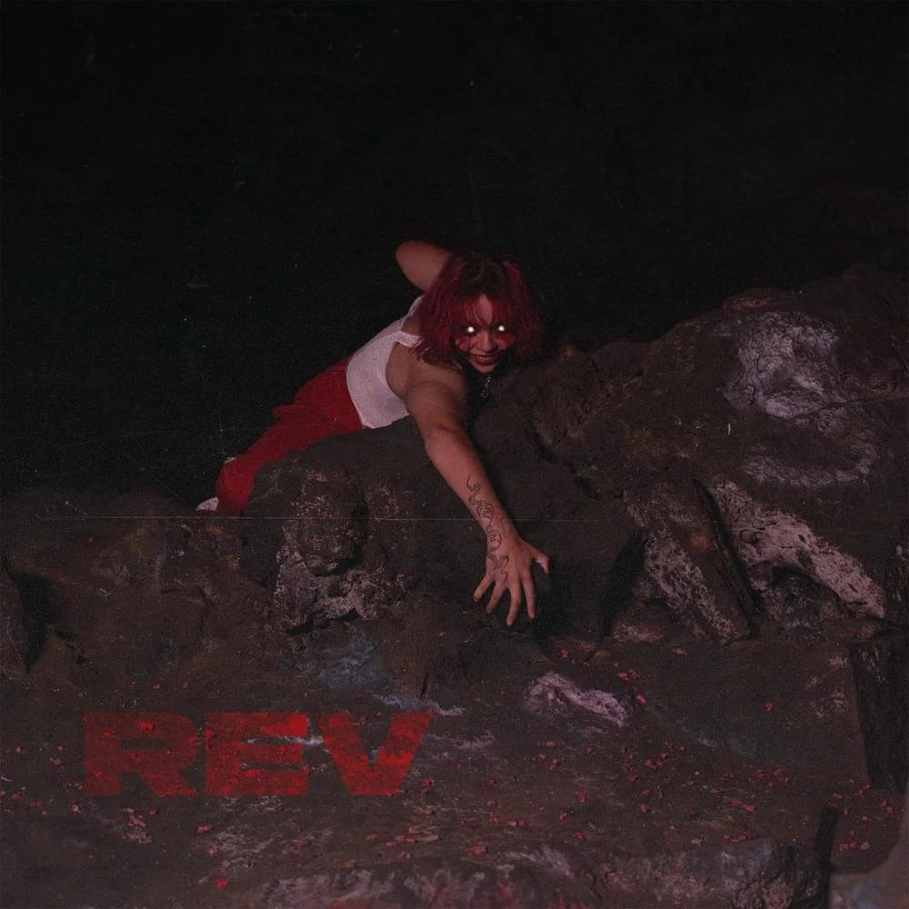 A person with red hair and glowing eyes climbing out of a pit of dark rocks in a dark environment with the word 'REV' in red on the rocks.
