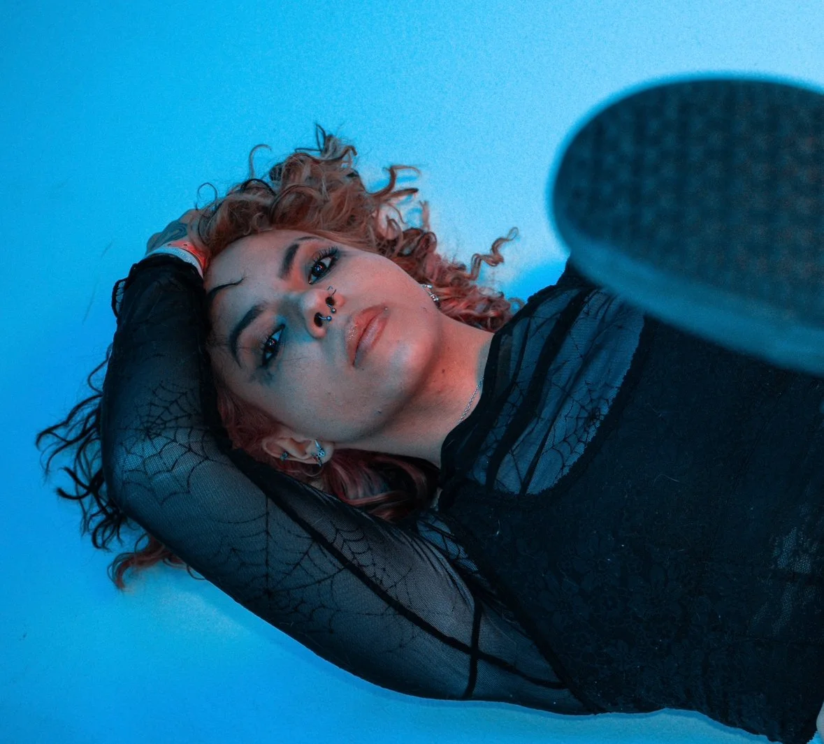 A young woman with curly hair and multiple facial piercings lying on her back, wearing a black lace top and fishnet sleeves, against a blue background.