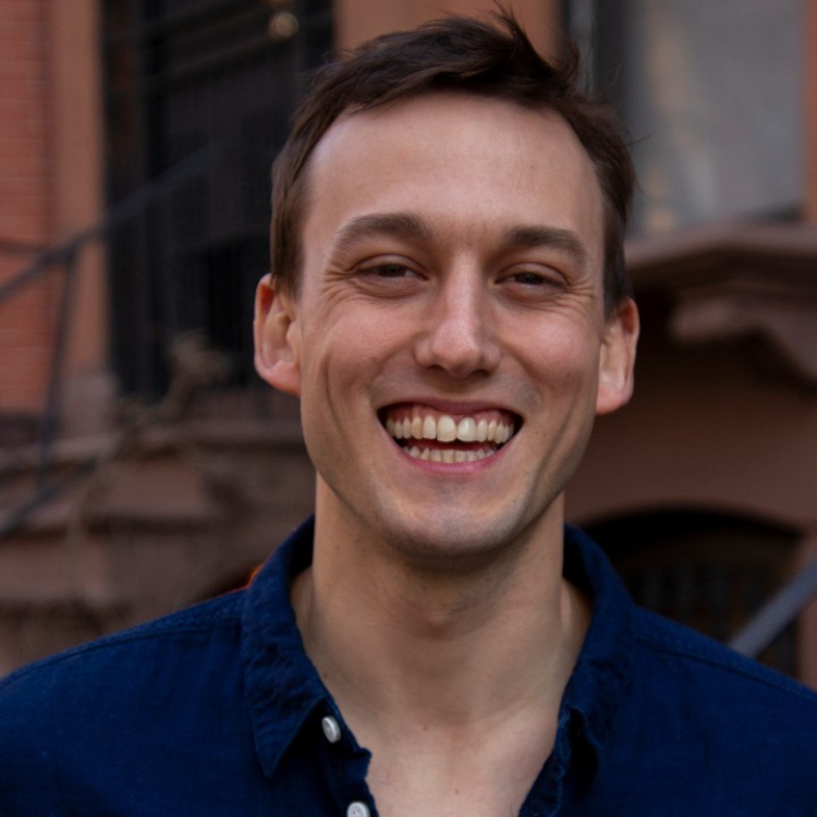 A young man with short brown hair smiling outdoors in front of an urban setting with a brick building and a staircase.