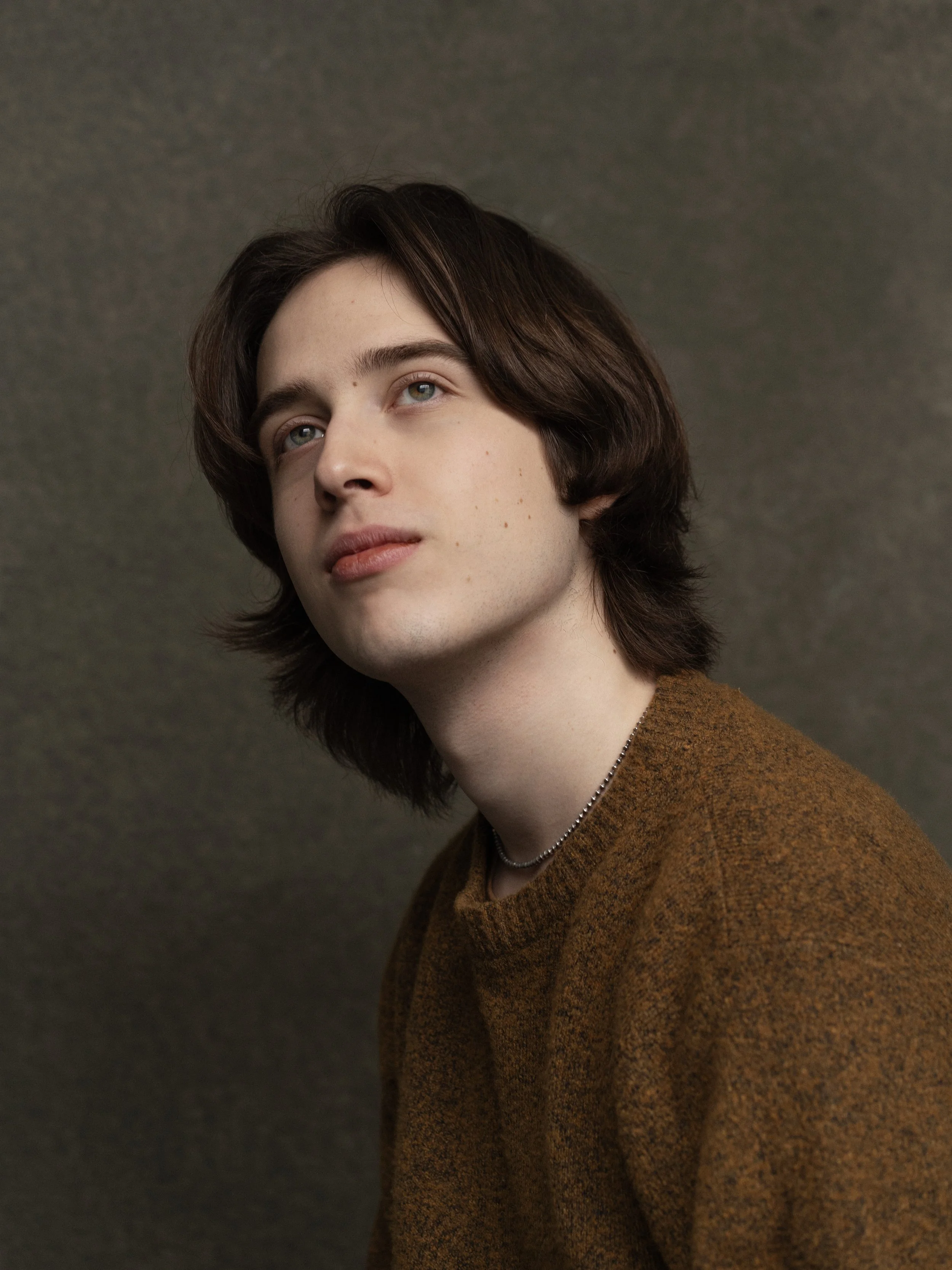 A young man with shoulder-length brown hair and blue eyes gazes slightly upward, wearing a brown sweater and a silver necklace, against a plain gray background.