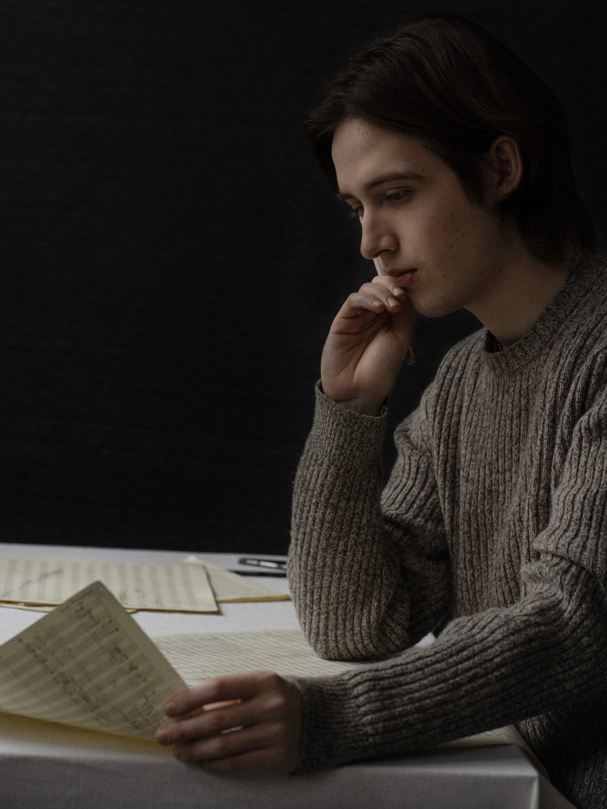 Yianni Frantzeskakis in a gray sweater reading sheet music at a white table, with a black background.