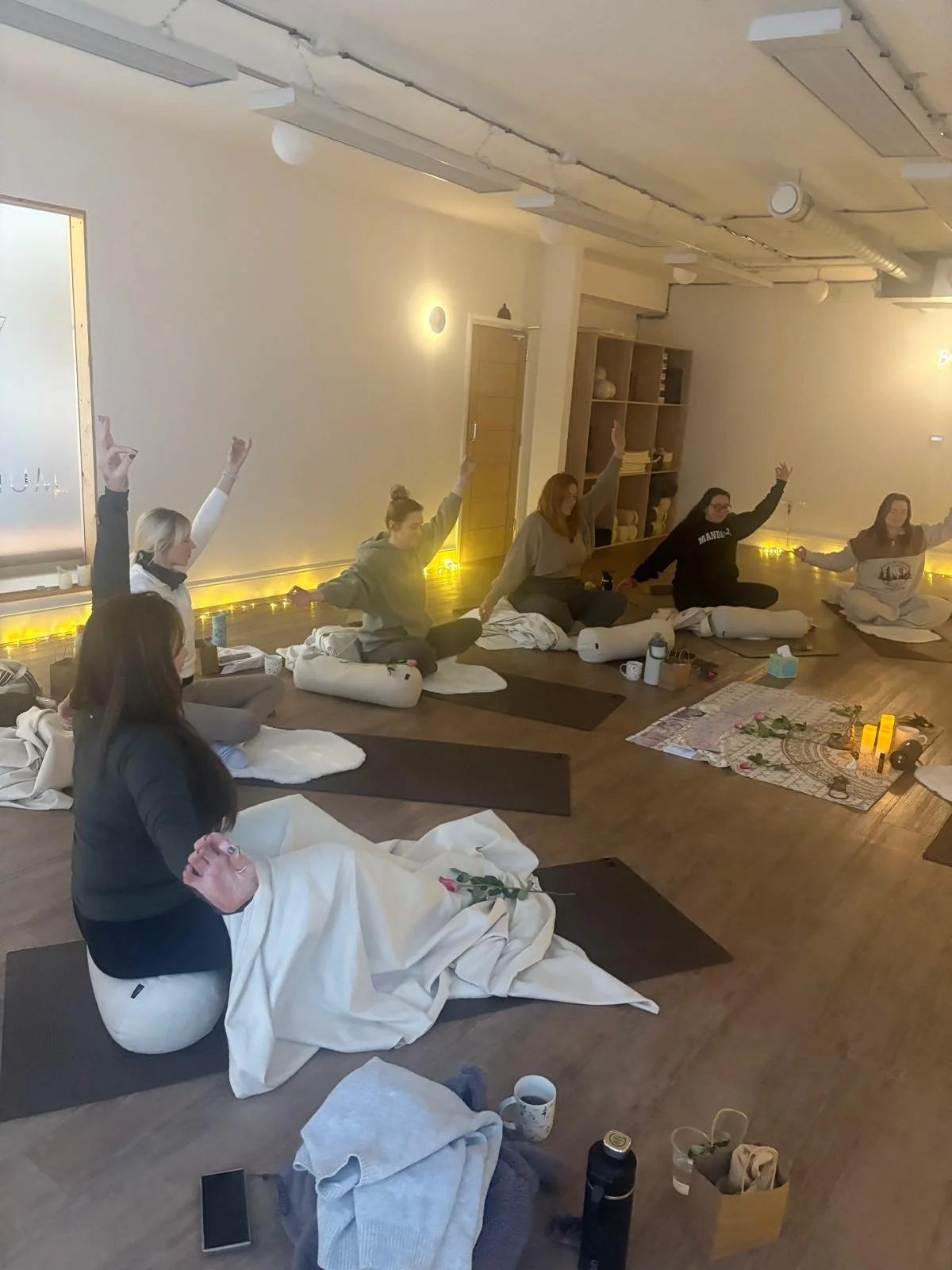 A group of women participating in a yoga or meditation class, sitting cross-legged on mats with blankets, some raising their hands, in a softly lit, cozy room with wooden floors and shelves.