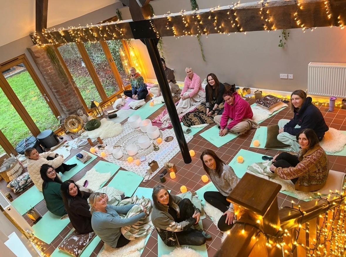 People sitting on yoga mats and cushions in a cozy, warmly lit room with string lights, large windows showing greenery outside, and decorative candles on the floor and table, participating in a group wellness or meditation session.