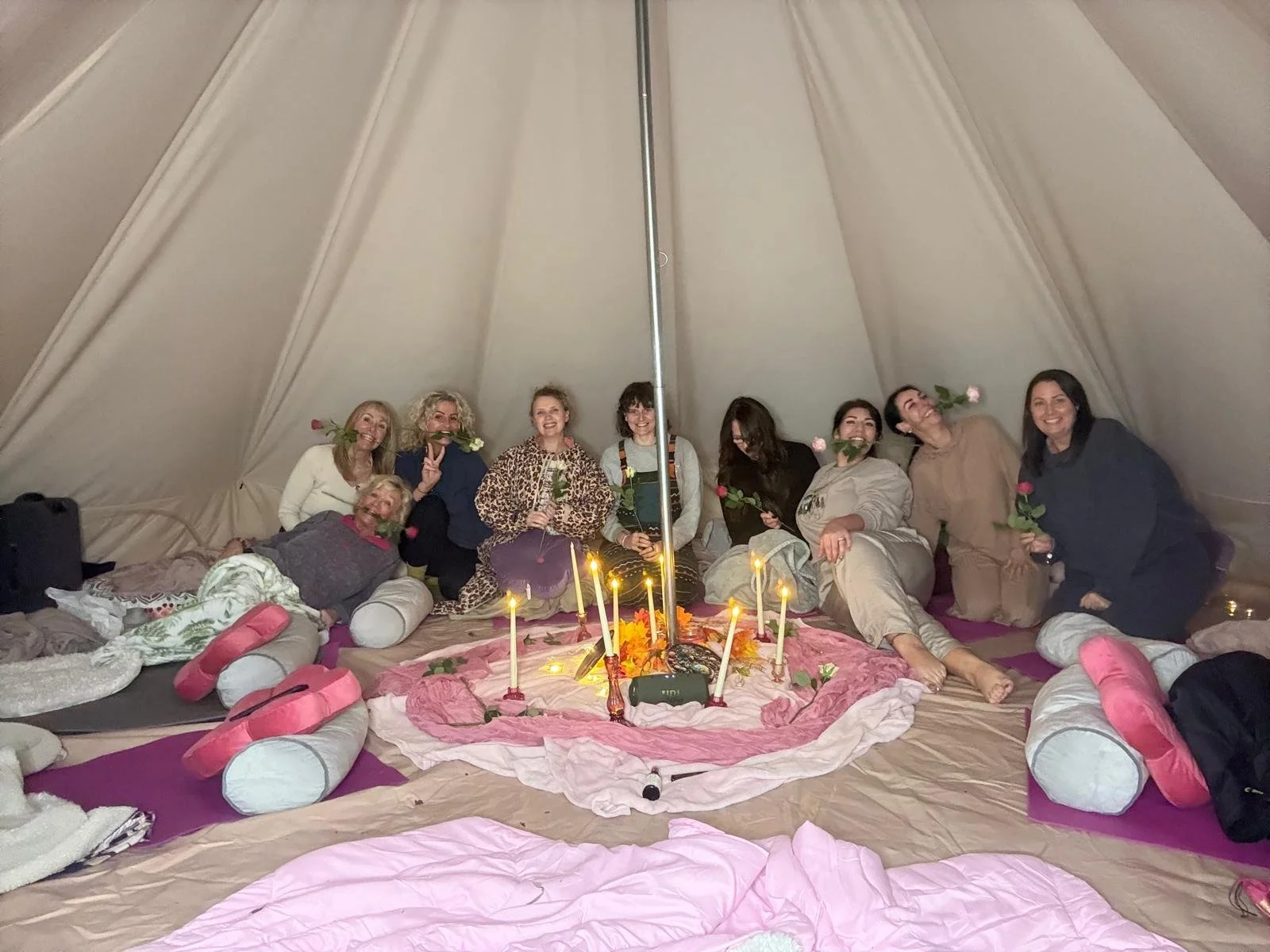 Group of women inside a large tent, sitting and lying on the floor around a pink cloth with candles, flowers, and small cushions, posing for a photo. Kundalini Yoga. Auric Alchemist retreat