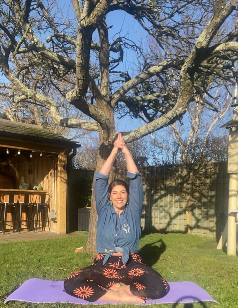 Woman practicing yoga outdoors on a purple mat, sitting cross-legged, raising her arms above her head, smile, in a backyard with a leafless tree and a wooden shed.
