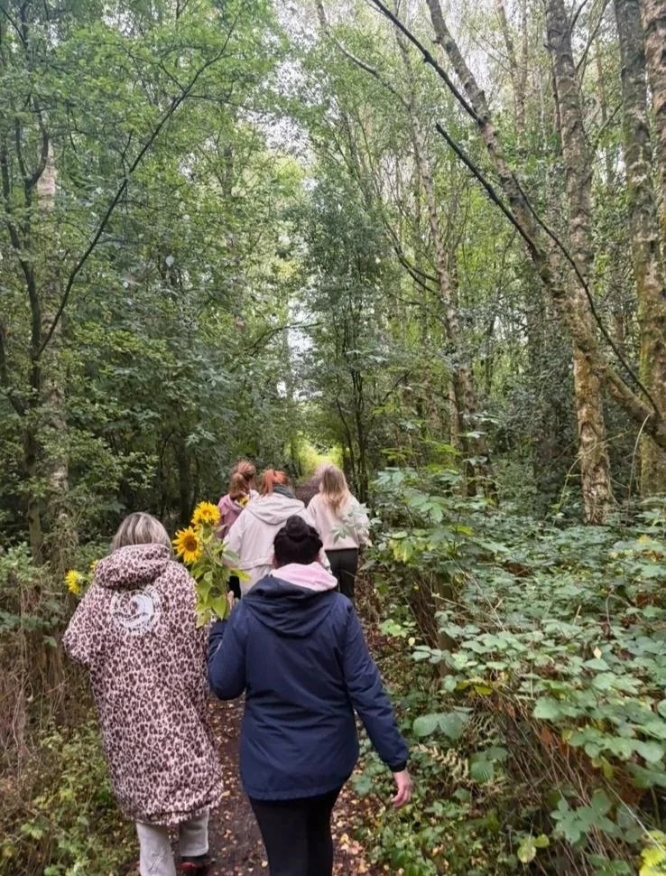 A group of people walking through a wooded trail, one person is holding a bouquet of yellow sunflowers, surrounded by green trees and foliage.
