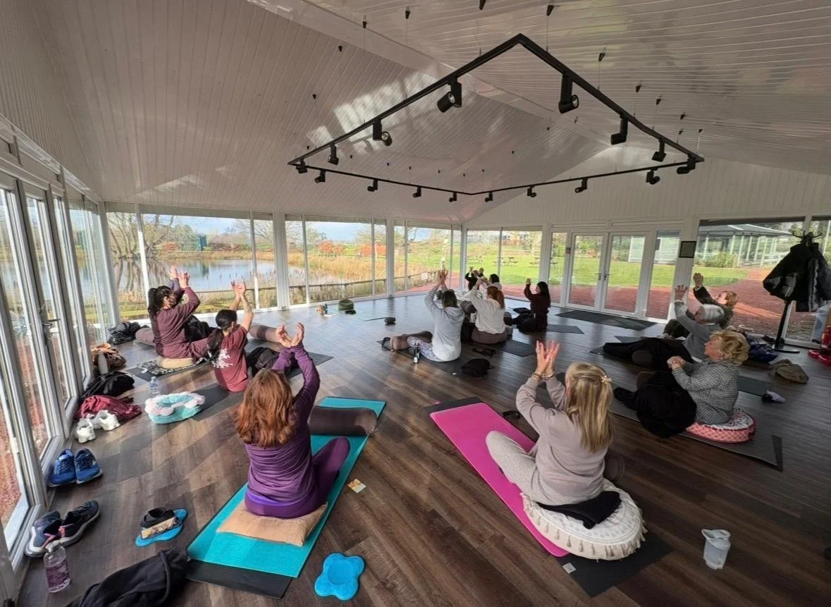 Group of people participating in a yoga or meditation class in a bright, spacious room with large windows overlooking a lake and outdoor scenery.