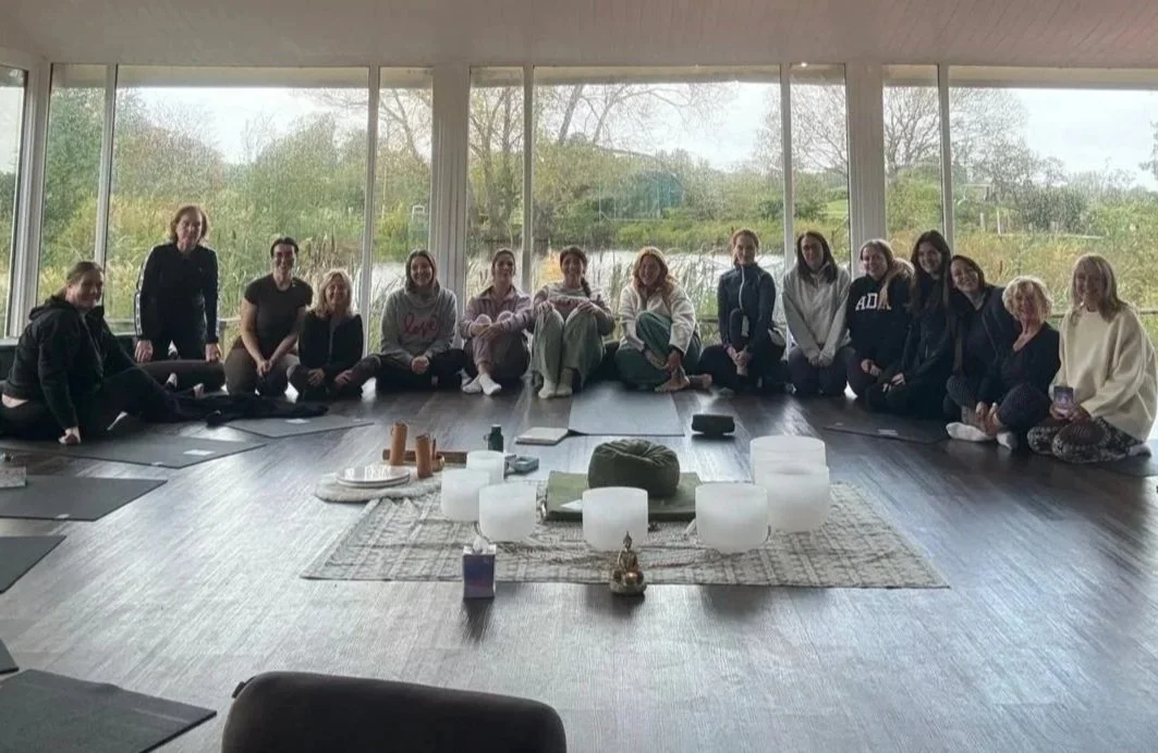 A group of people sitting in a circle on the floor of a spacious room with large windows and a view of trees outside. They are seated around a decorated area with candles and pillows, likely for a meditation or yoga session.