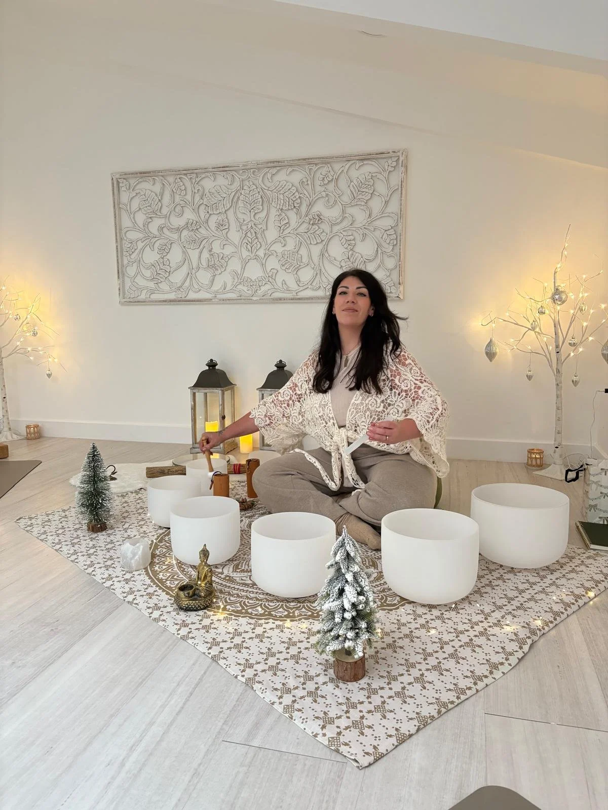 Woman sitting cross-legged on a patterned rug amid holiday decorations, including small Christmas trees, lanterns, and white bowls, with lights and ornamental trees in a cozy, well-lit room.