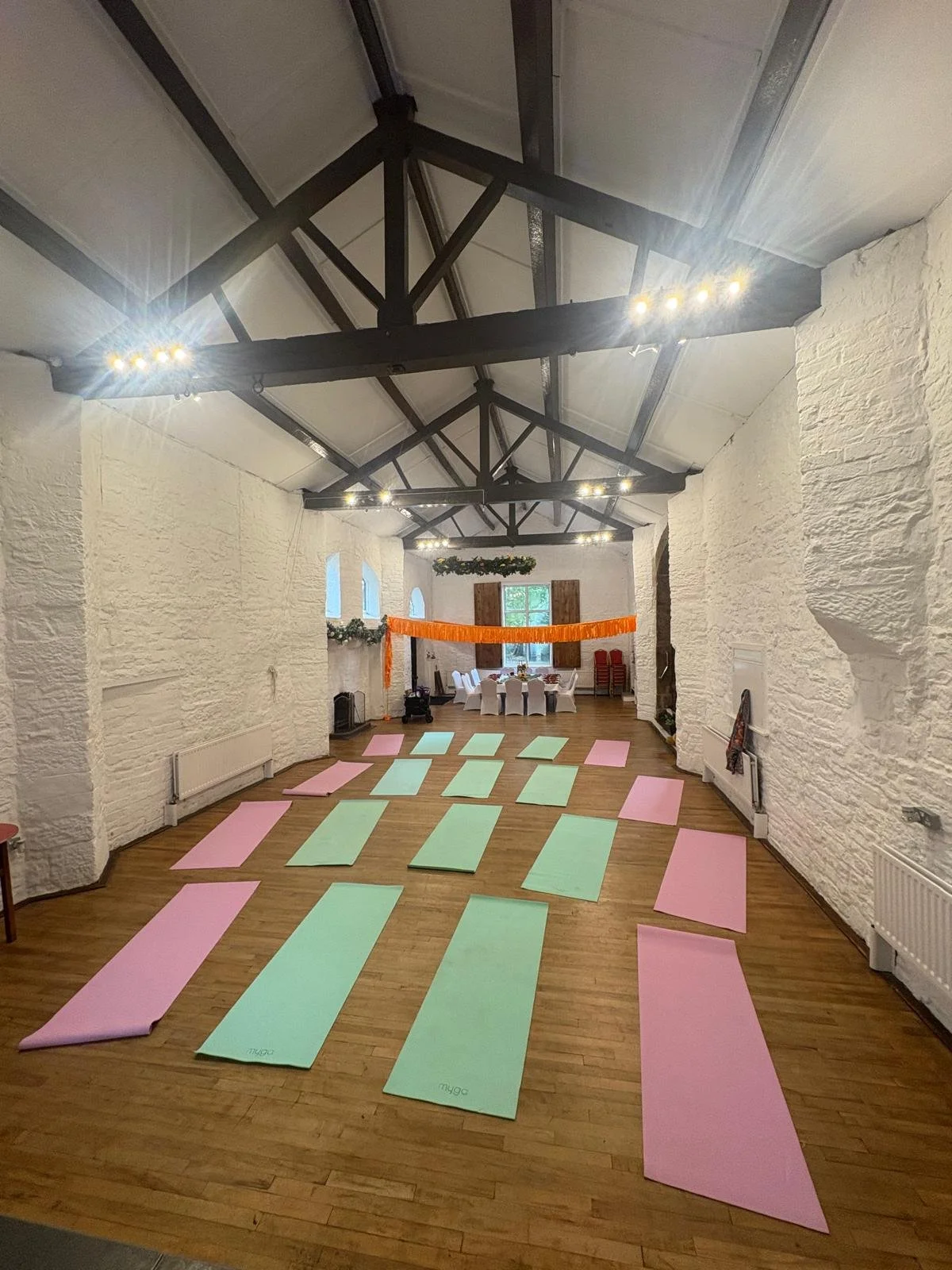 A spacious room decorated for a yoga or wellness event with pink and green mats laid out on wooden floor, white brick walls, high ceiling with black beams and string lights, and tables and chairs at the far end. Kundalini Yoga. Auric Alchemist
