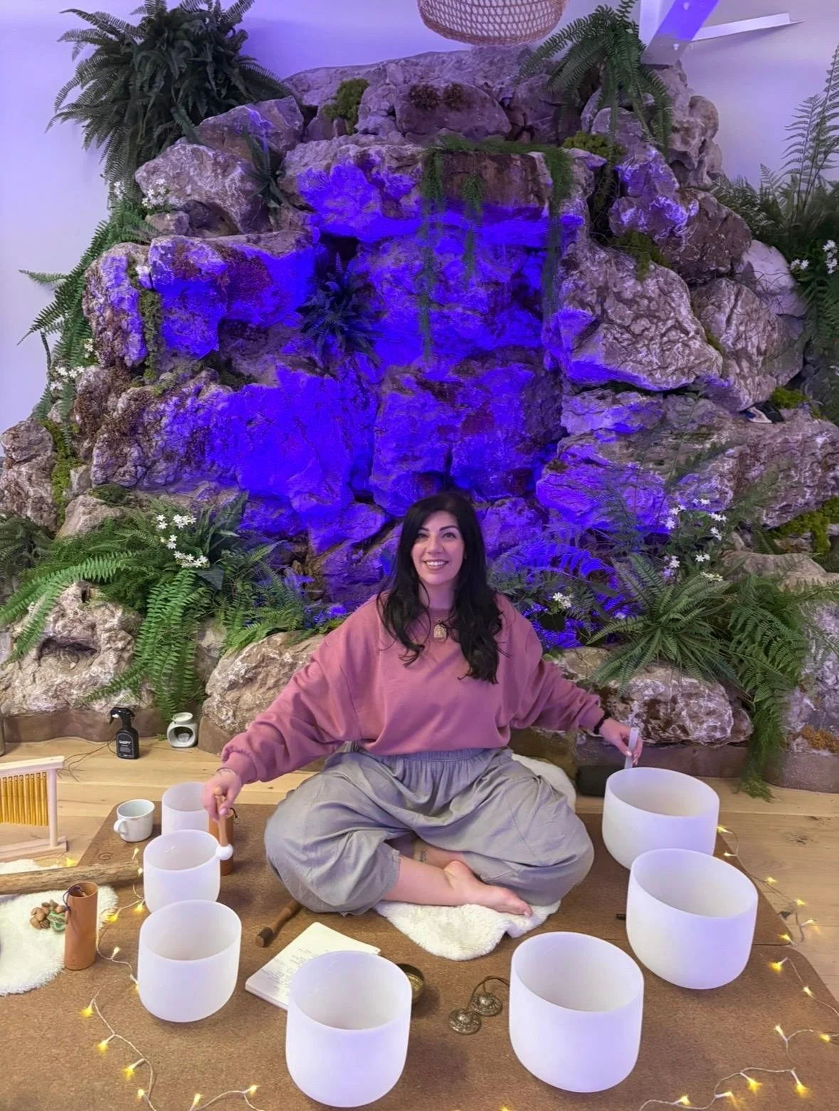 A woman sitting cross-legged on a rug, surrounded by white singing bowls, with a decorative fountain and lush green plants with white flowers in the background.