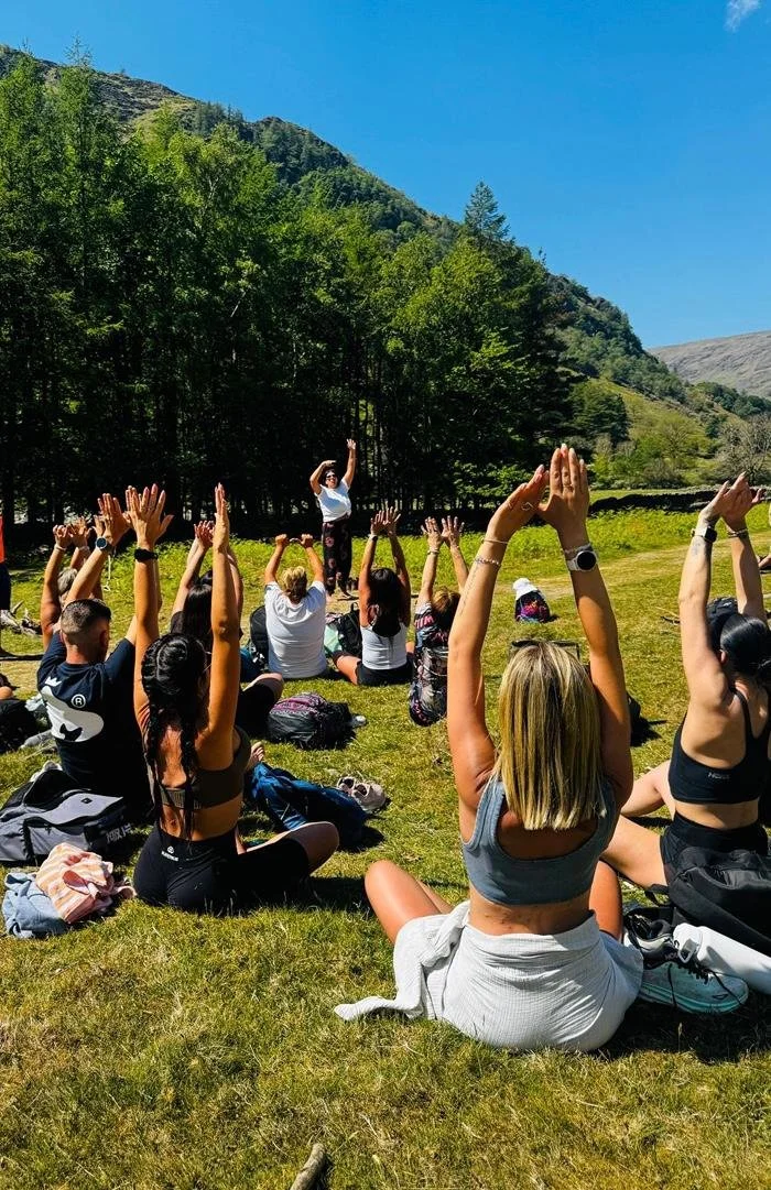 People participating in outdoor yoga or meditation session on a grassy field surrounded by trees and mountains on a sunny day.
