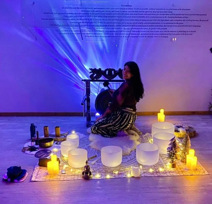 A woman sitting on a white fur rug amidst candles and decorative items, with a projection of blue light beams and text on the wall behind her, creating a calming atmosphere.