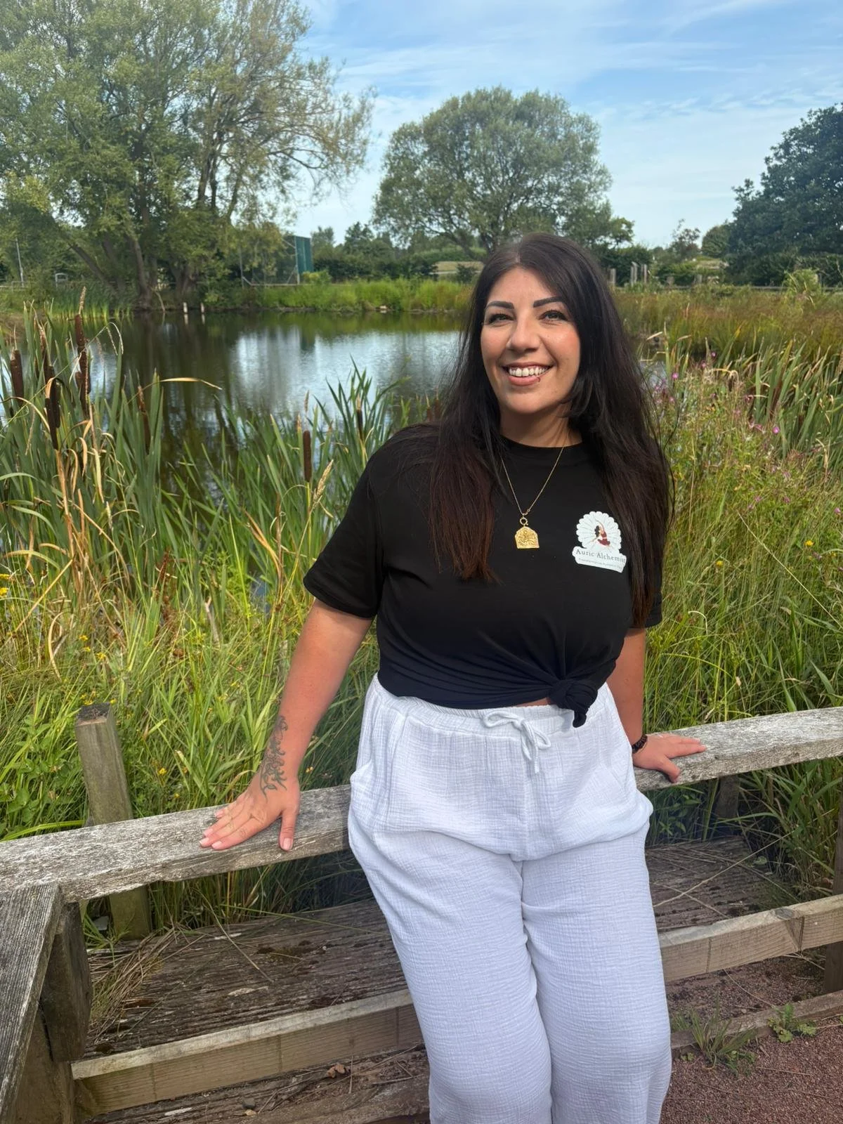 Smiling woman with dark hair, wearing a black t-shirt and white pants, standing outdoors by a wooden fence near a pond with tall grass and trees in the background.
