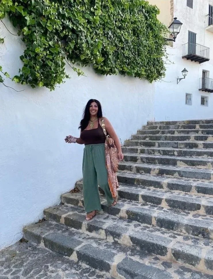 A woman smiling and standing on stone steps next to a white wall with green ivy above. She is holding a phone, wearing a brown top, wide green pants, and sandals, with a patterned scarf and a shoulder bag. There are small balconies and lanterns on the building in the background.