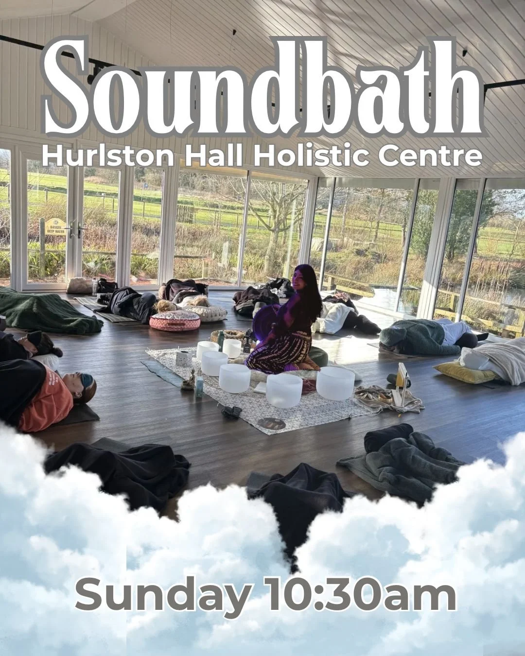 People participating in a soundbath meditation session inside a bright room with large windows overlooking a scenic outdoor landscape, seated or lying on mats and cushions, with a woman playing singing bowls on a central rug.