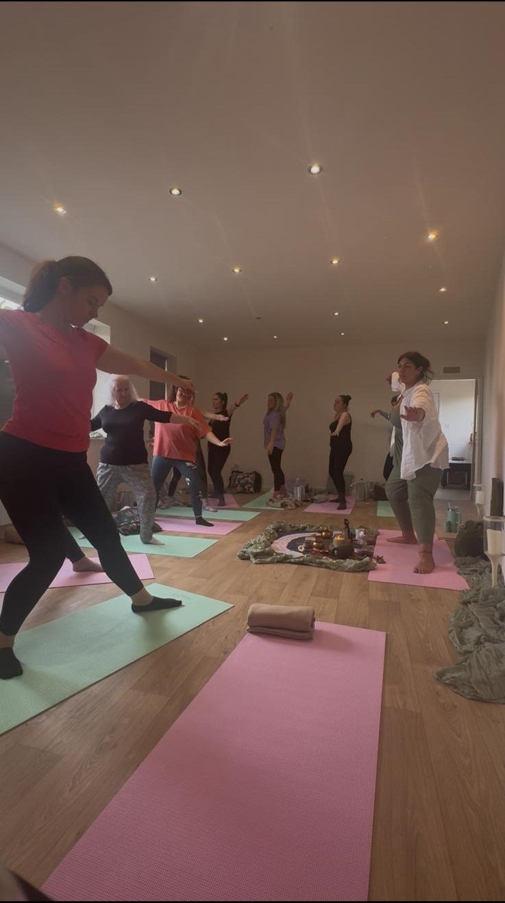 A group of women participating in a yoga or meditation class on pink and green mats in a spacious room with a wooden floor and ceiling lights. Some women are standing on one leg with arms outstretched, practicing poses or balancing.