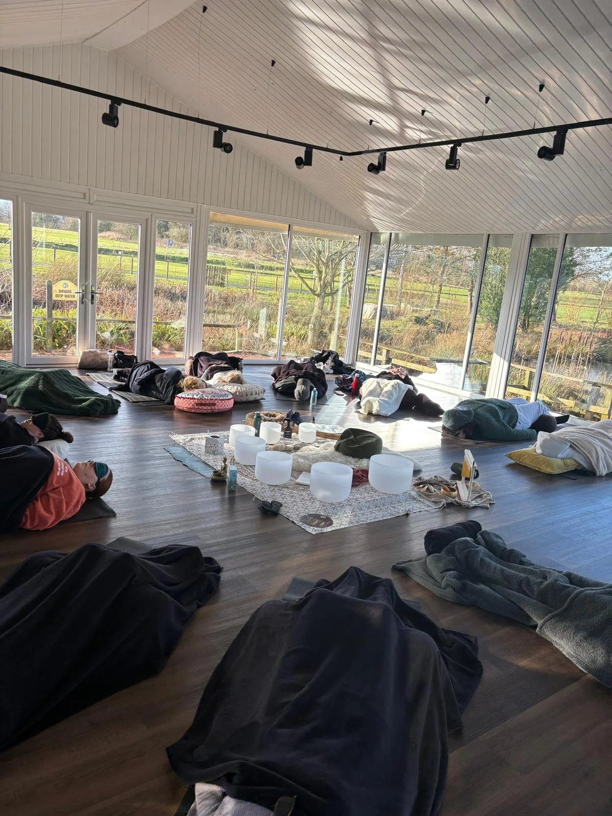 People lying on the floor in a bright yoga or meditation room with large windows overlooking a garden, with a central music setup including crystal bowls and candles.