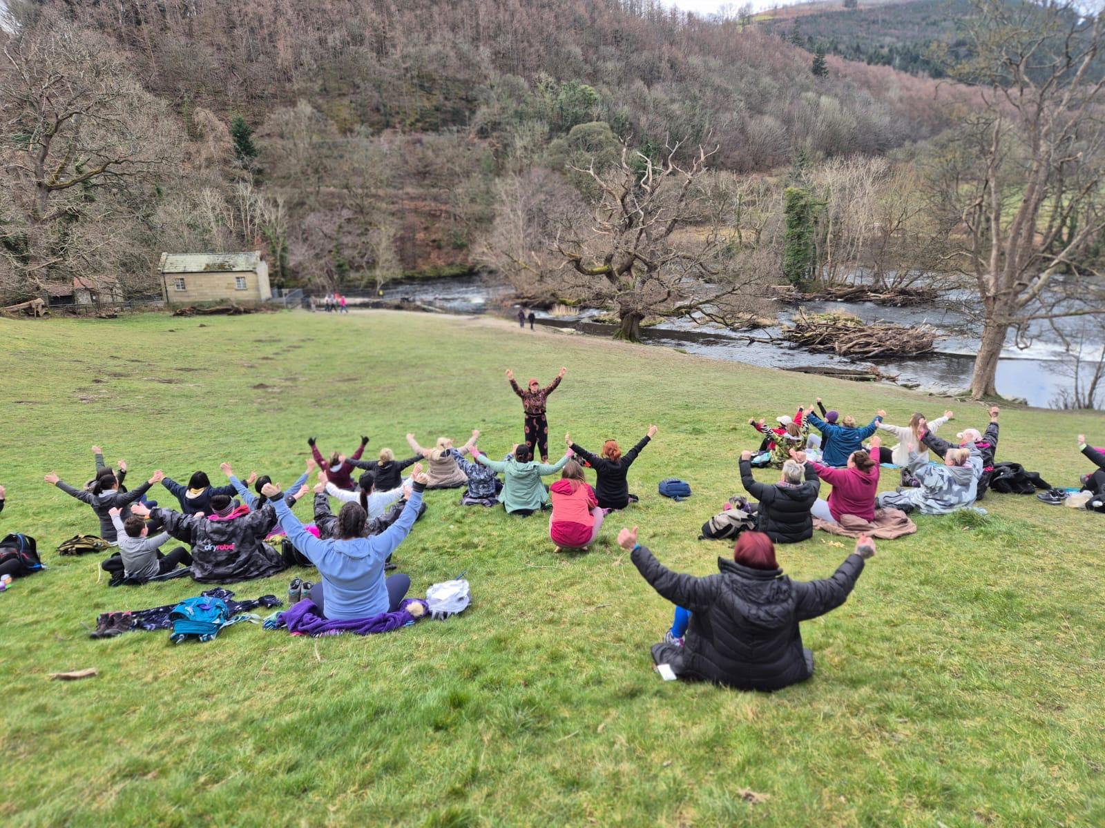 Group of people practicing yoga or meditation outdoors on a grassy field near a river, with trees and hills in the background.