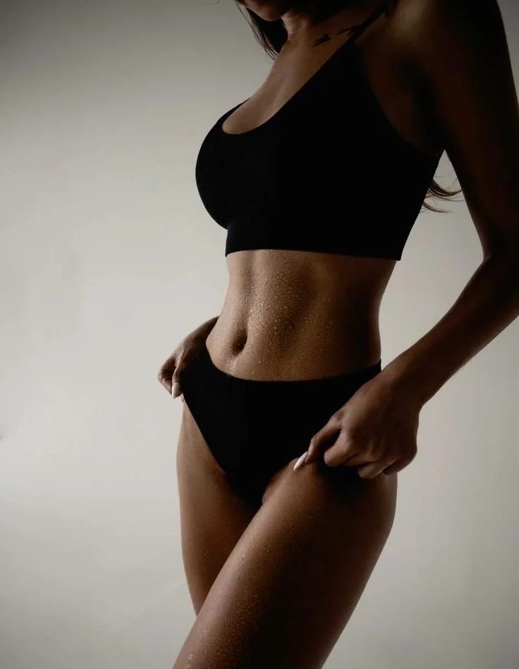 Close-up of a woman's midsection, wearing black sports bra and underwear, with a neutral background.