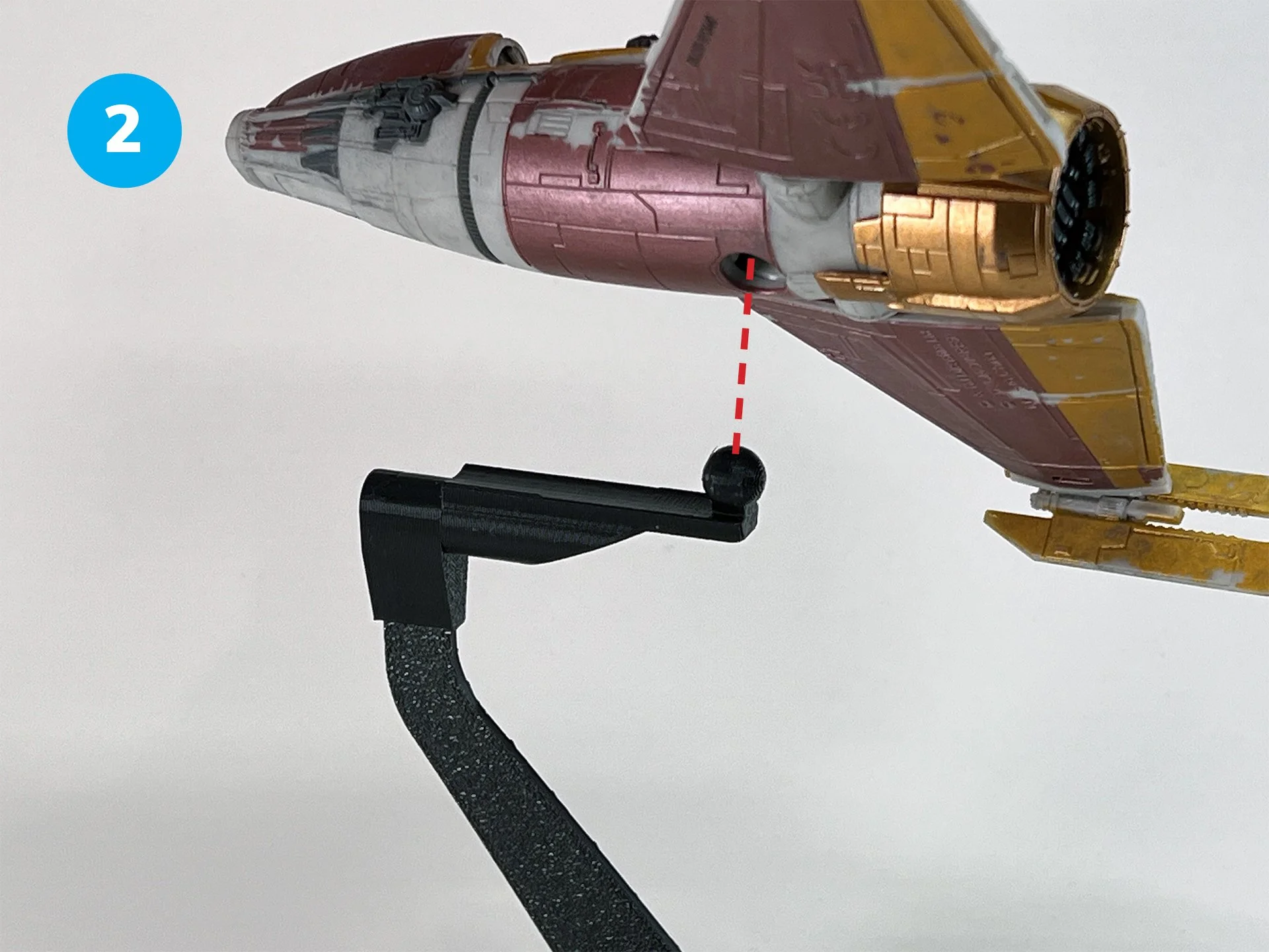 Close-up of a plastic model of a spacecraft or fighter jet being mounted on a stand, with a focus on the nose cone and cockpit area. The model features red, gray, and gold colors, with a black mounting arm attached.