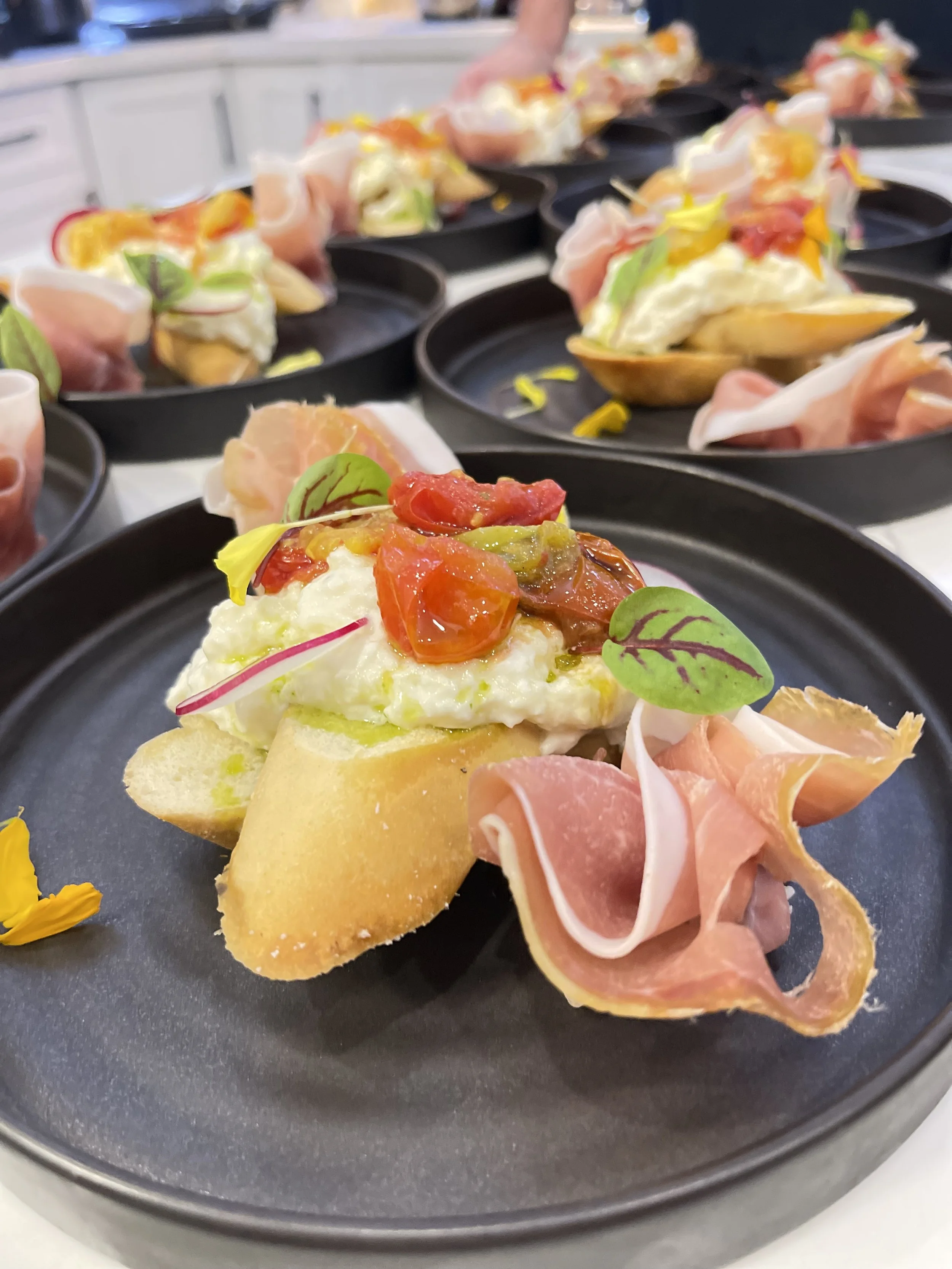 Close-up of a plate with bread, feta cheese, cherry tomatoes, microgreens, and prosciutto, with multiple similar plates in the background.