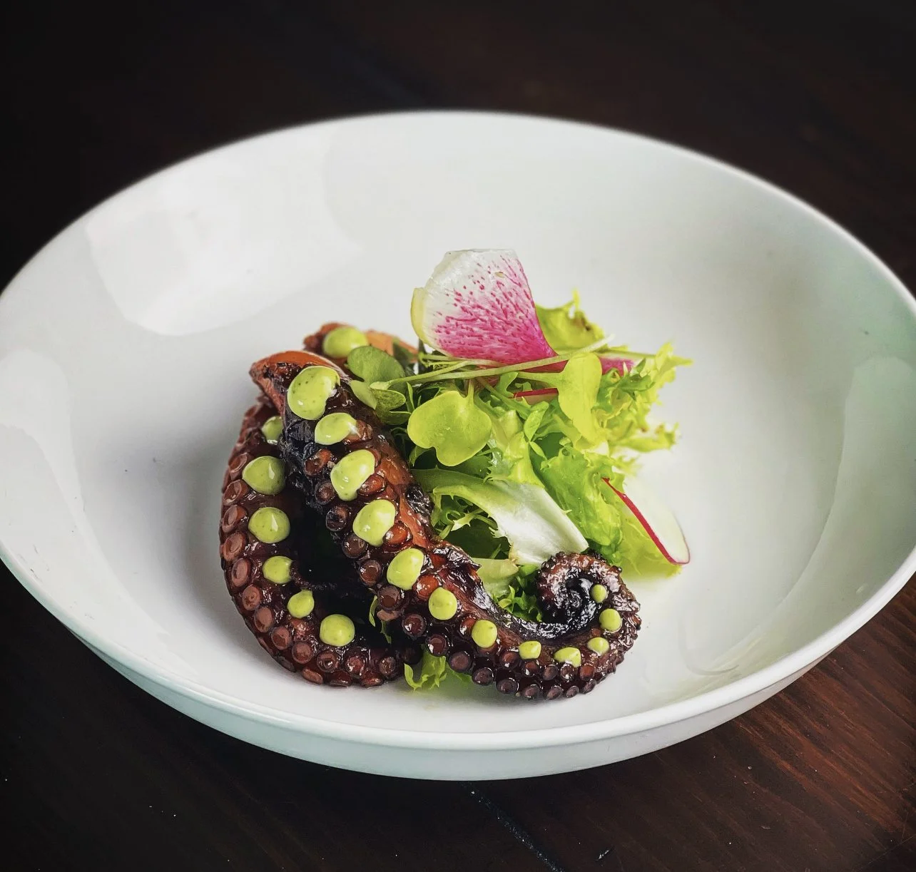 Grilled octopus tentacle served on a white plate with microgreens and edible flower