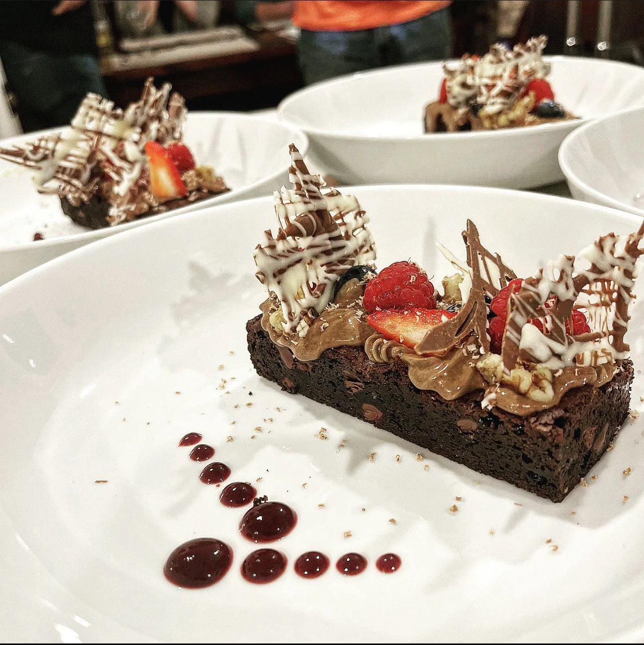 A slice of chocolate cake garnished with strawberries, raspberries, and decorative chocolate pieces on a white plate with raspberry sauce dots.