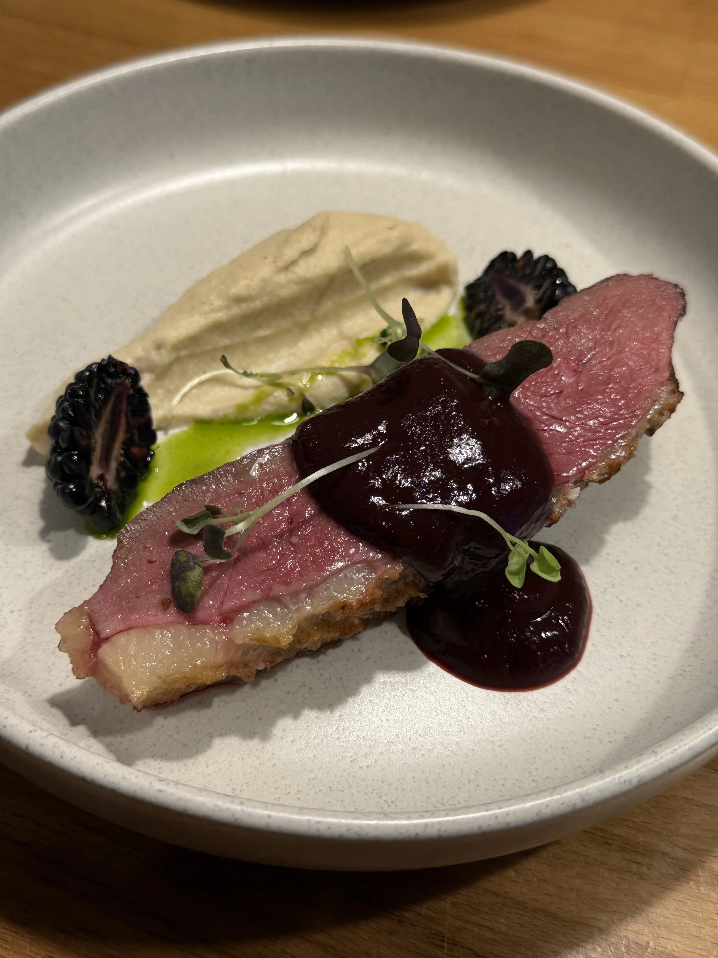 Slices of cooked meat with dark berry sauce, a dollop of creamy mashed potatoes, blackberries, microgreens, and green oil on a white plate. duck breast