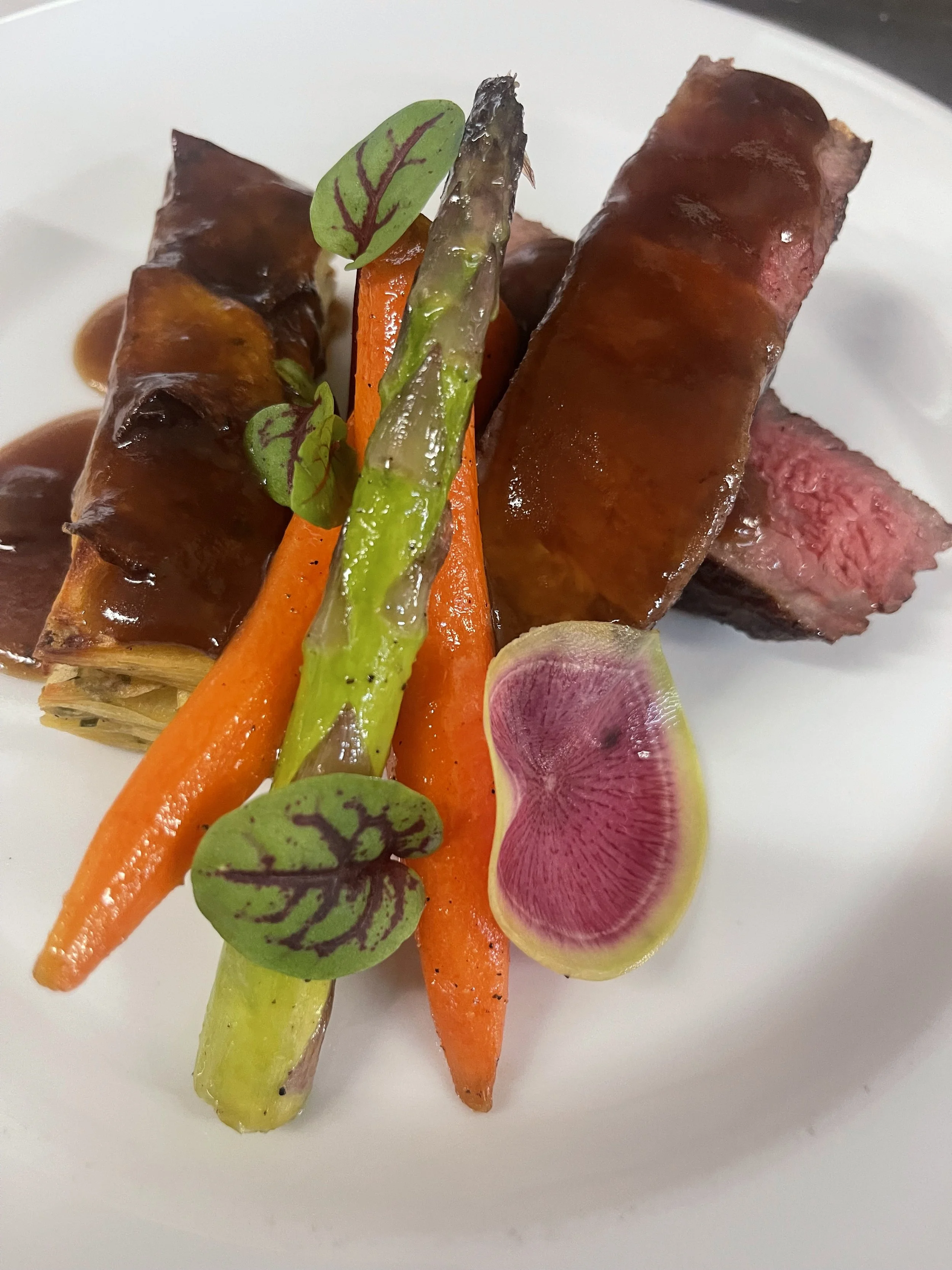 A plated dish with slices of medium rare steak, glazed carrots, asparagus, radish, and microgreens. strip loin. private chef