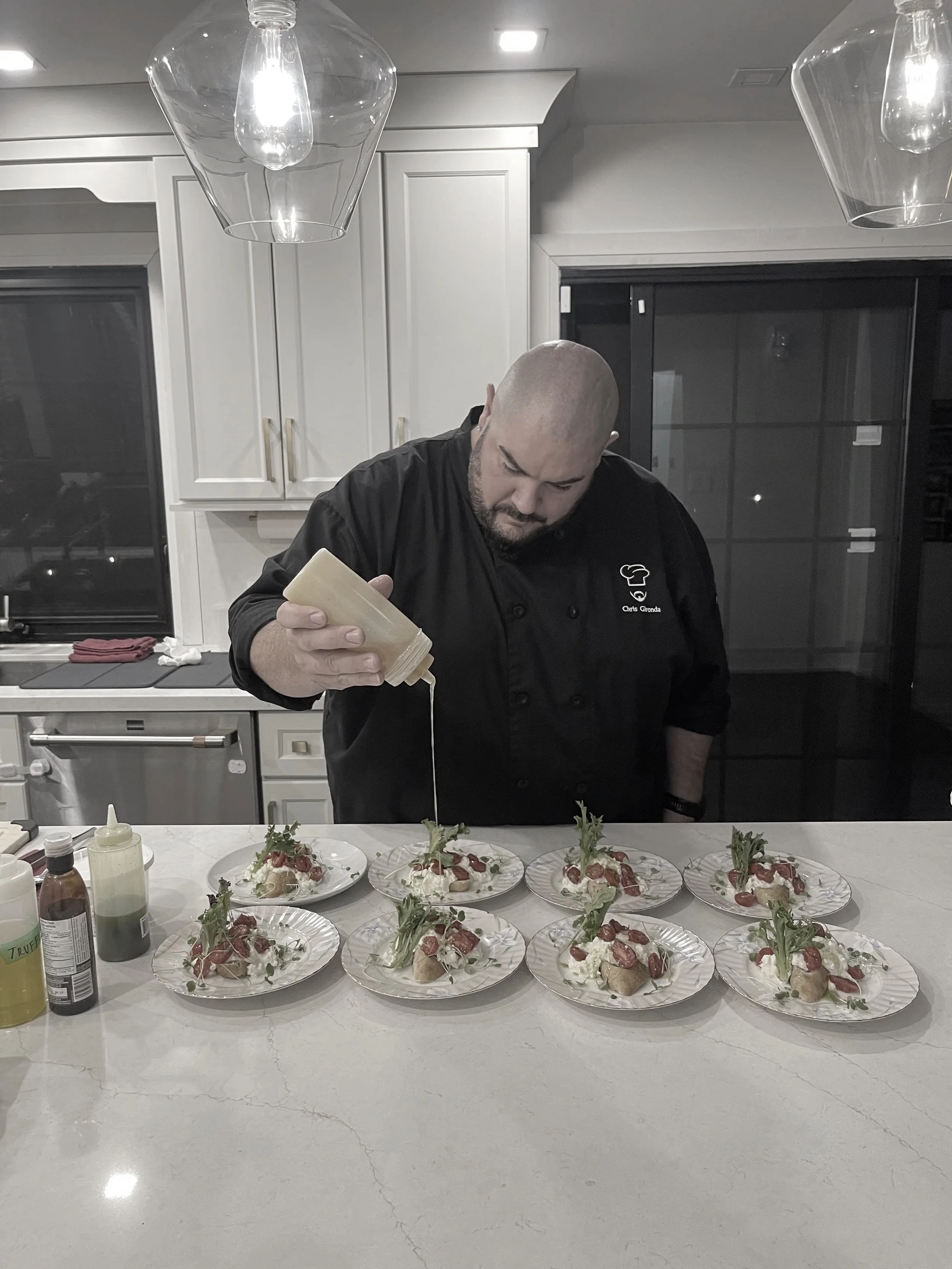 A chef in a black uniform is pouring sauce over several plates of food arranged on a kitchen island. The plates contain a dish garnished with herbs and tomatoes.