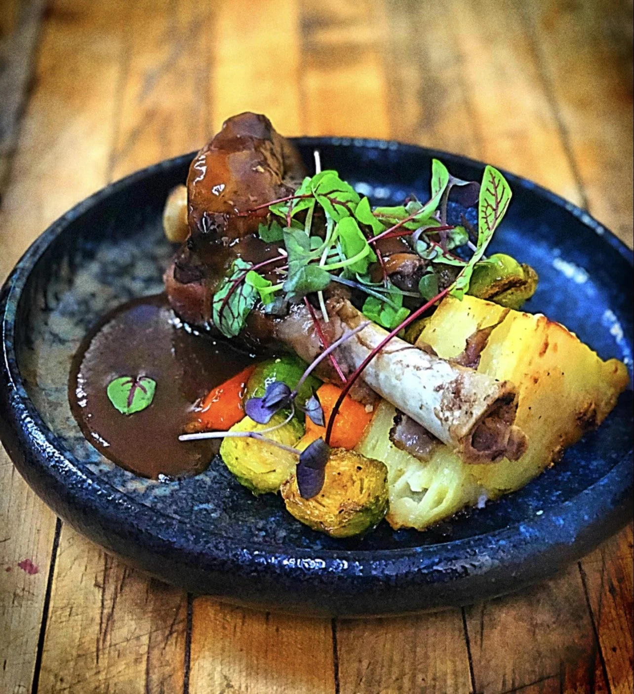 Close-up of a plate of roasted vegetables and meat stew with microgreens on top, served on a black ceramic dish on a wooden table. braised lamb shank