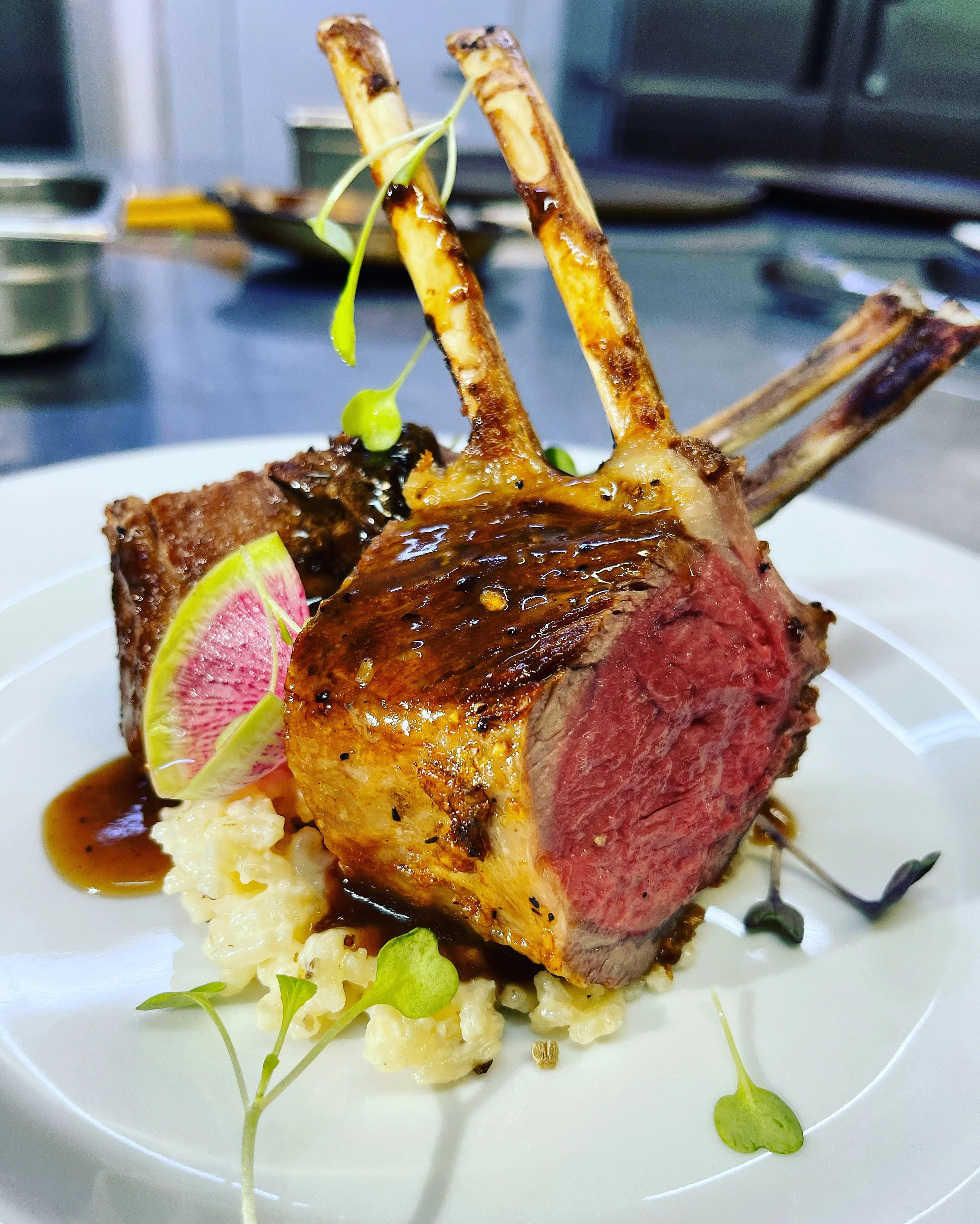 A cooked rack of lamb served on a white plate with mashed potatoes, pink radish slices, microgreens, and a dark sauce.