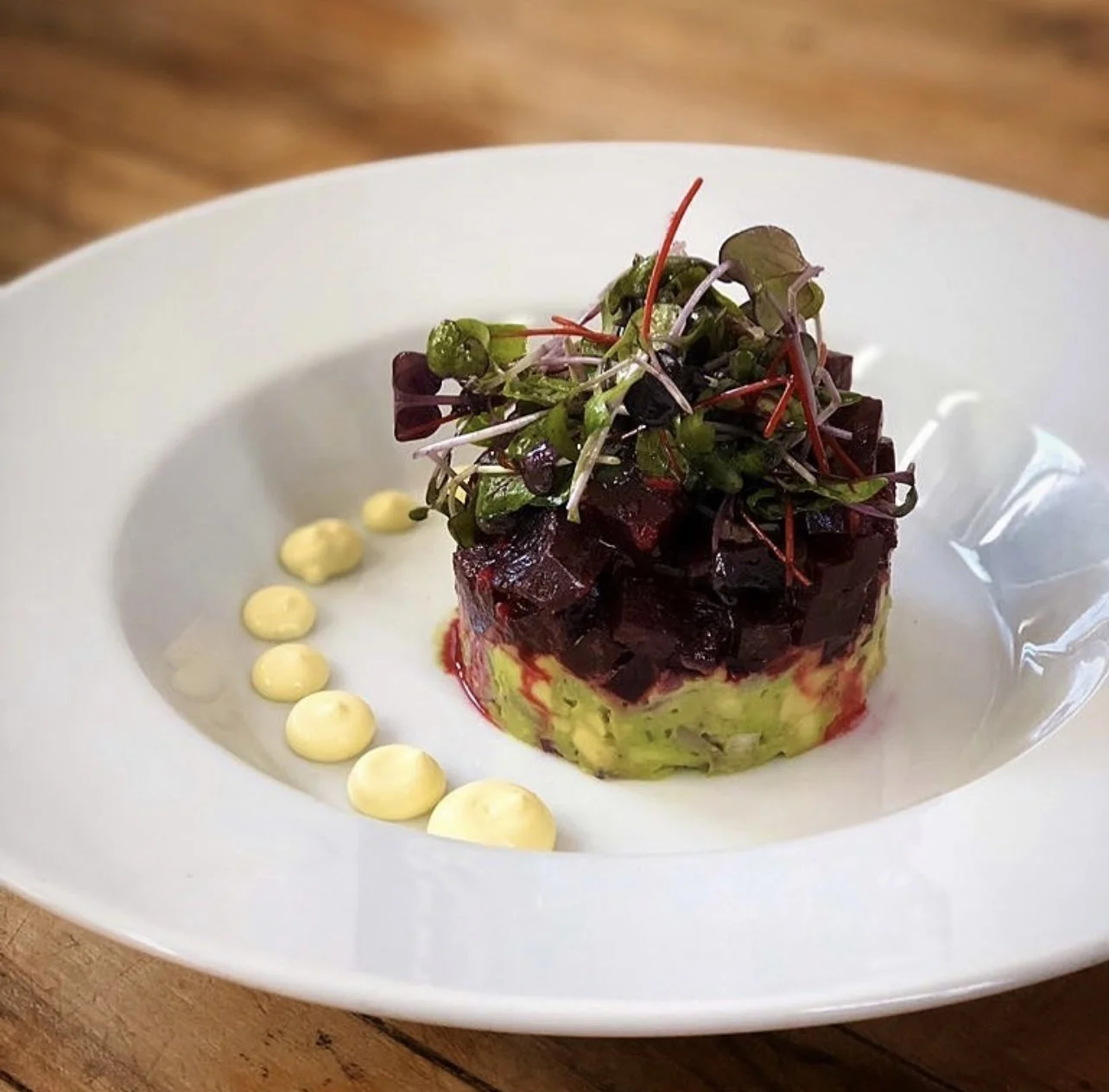 A plated dish featuring a cube of beet salad on top of a layer of mashed avocado, garnished with microgreens, and decorated with a pattern of mayonnaise dots on the side, served on a white plate.