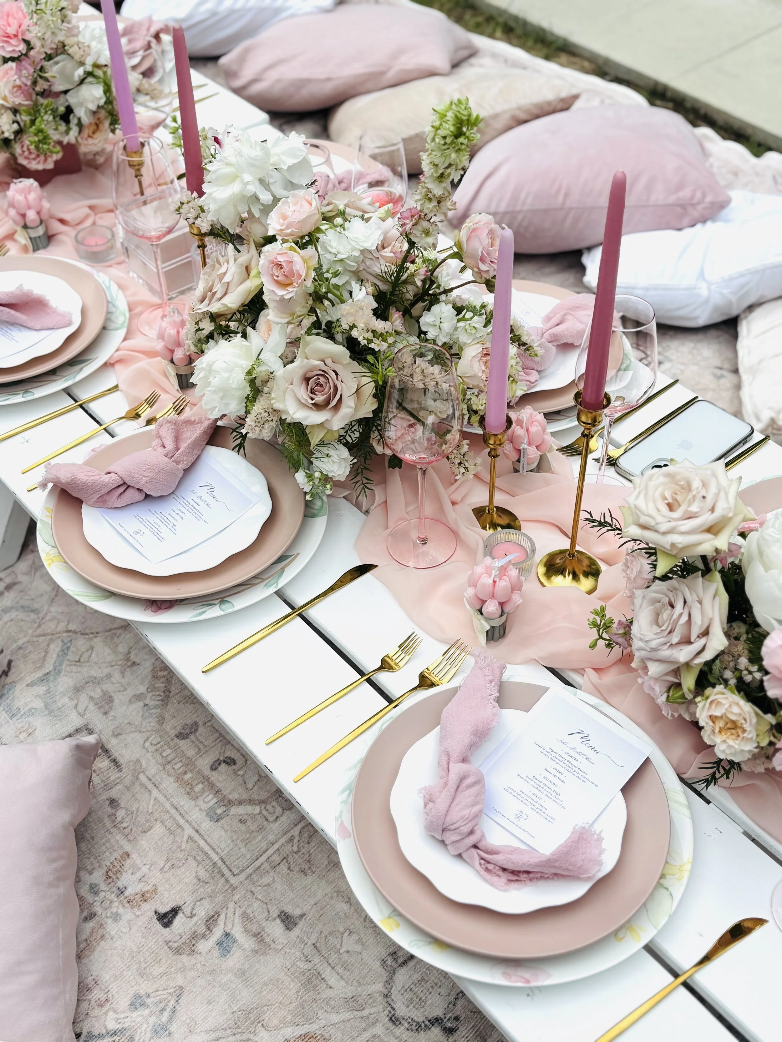 Elegant outdoor dining table decorated with pink and white flowers, pink candles, gold cutlery, pink napkins, and pink glassware, set on a white table with pillows and blankets in the background.