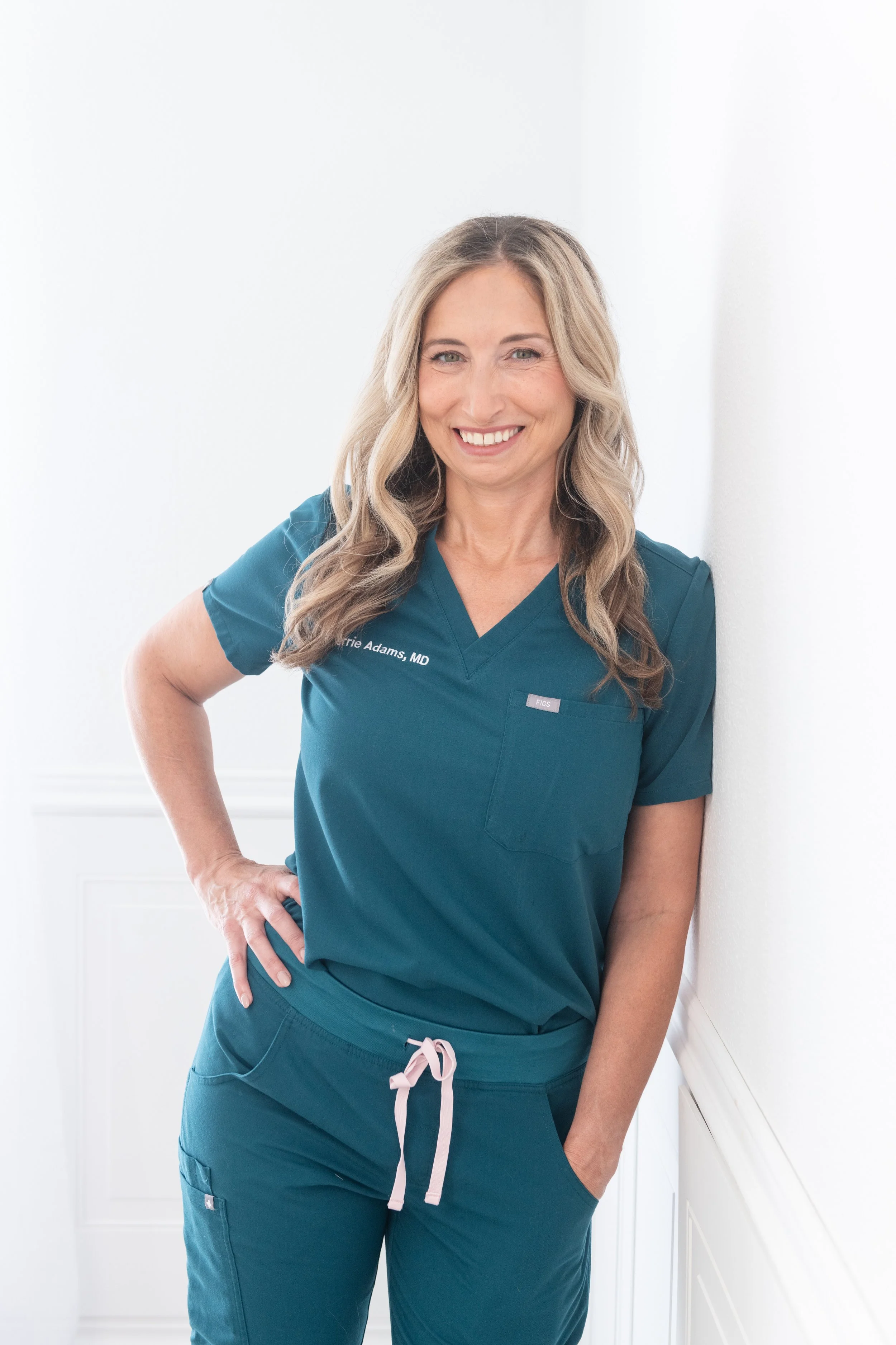 A woman in teal medical scrubs standing and leaning against a white wall, smiling at the camera.