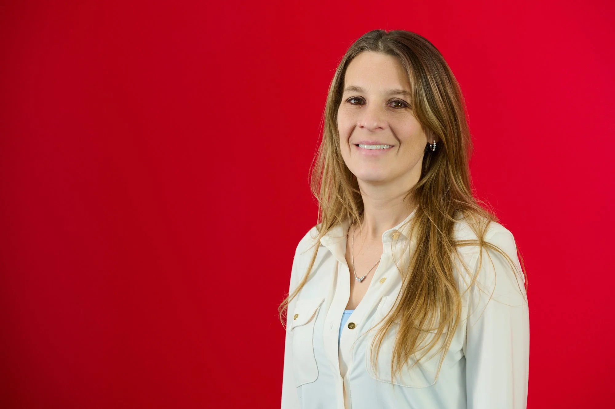 A woman with long light brown hair, wearing a white collared shirt and jewelry, smiling in front of a red background.