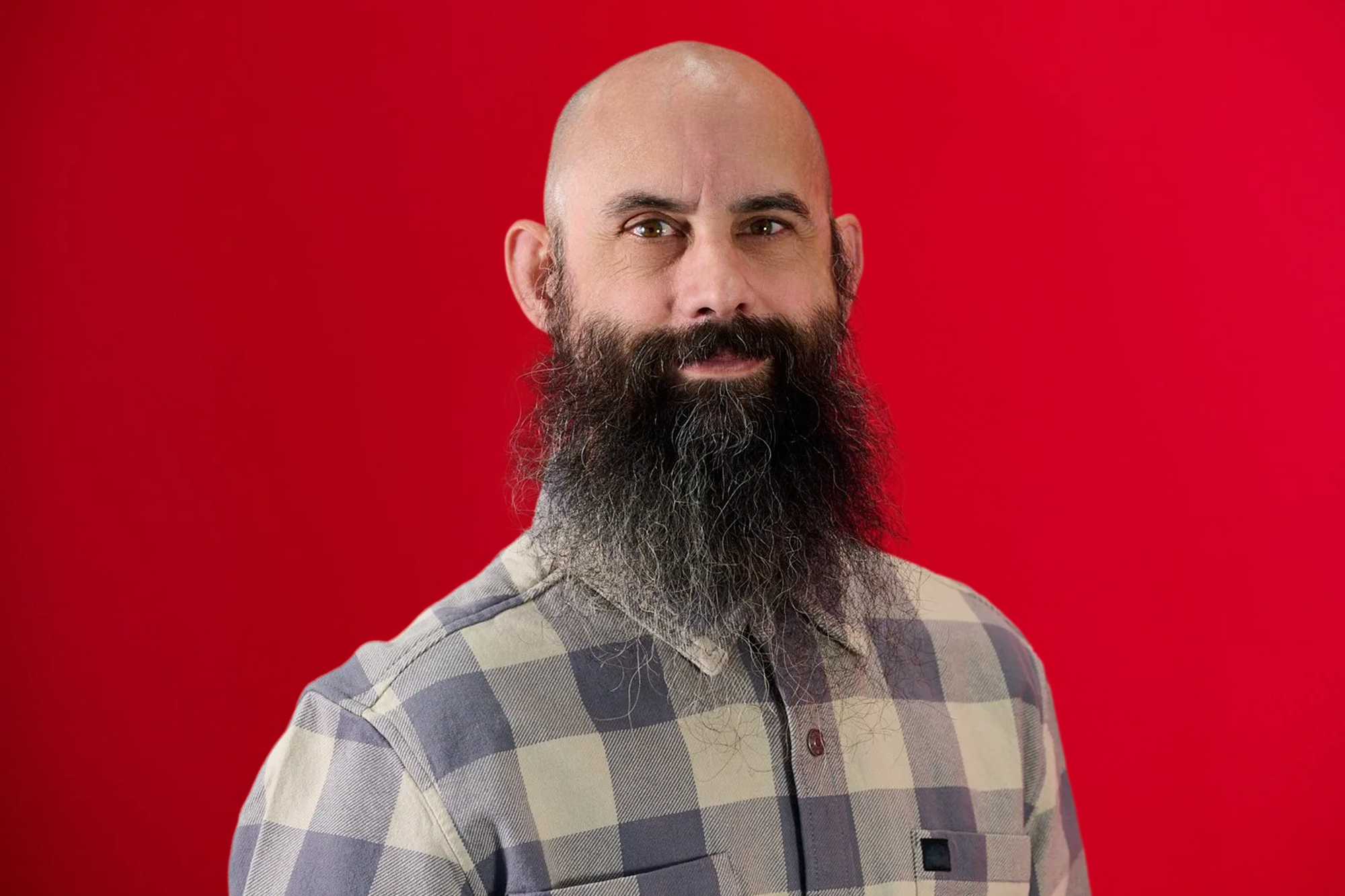 A man with a bald head and a full, long, bushy beard standing in front of a red background, wearing a plaid collared shirt.