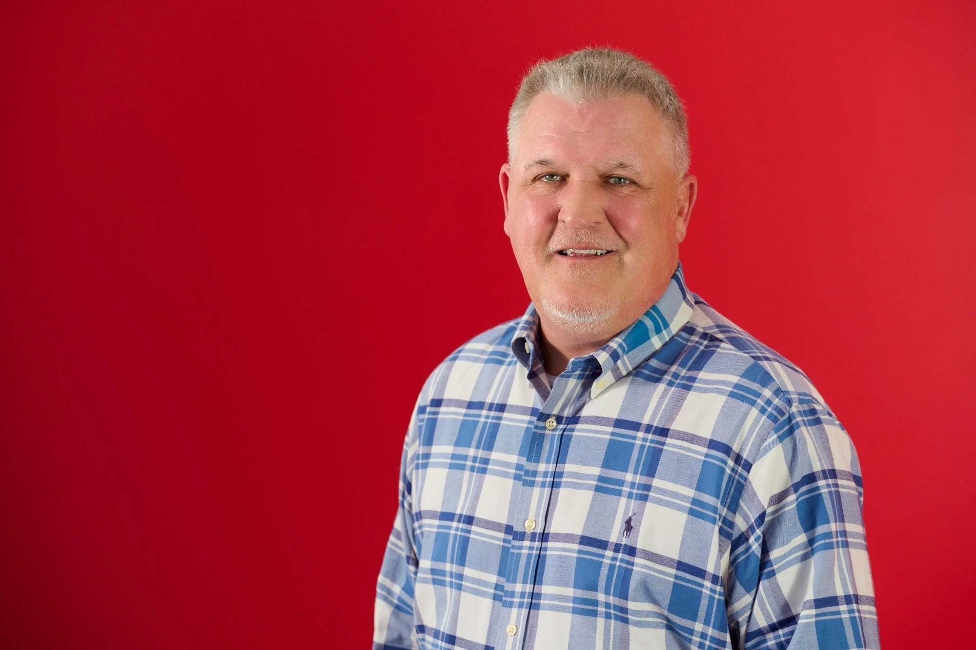 A middle-aged man with gray hair and a short gray beard smiling, wearing a blue and white plaid button-up shirt, standing against a red background.