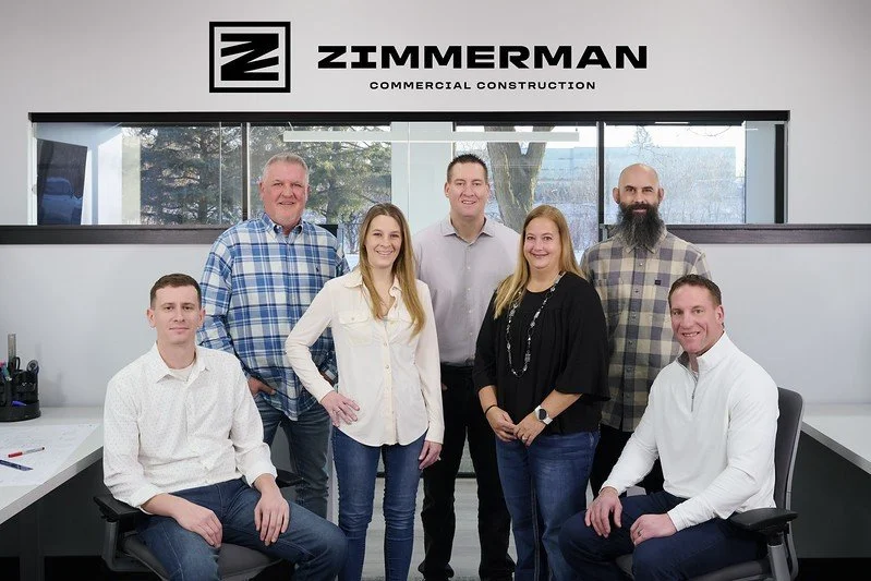 Group of seven professionals in office, with a sign reading 'Zimmerman Commercial Construction' behind them.