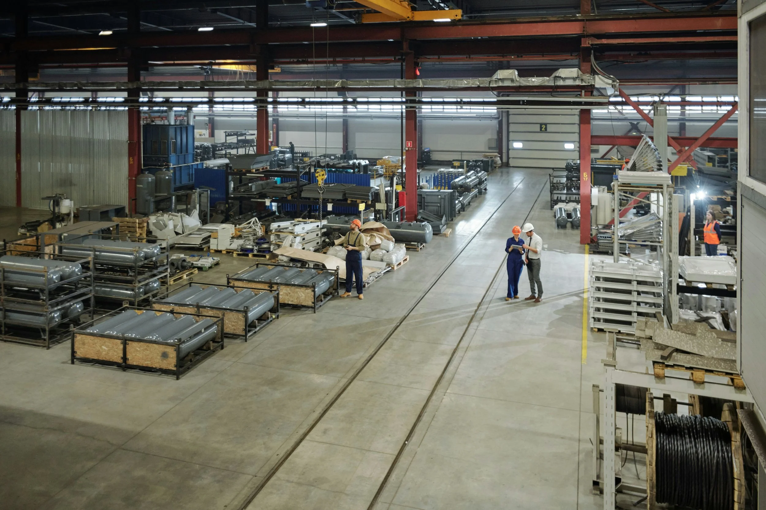 Inside a large industrial warehouse with workers wearing safety helmets and vests, surrounded by metal parts, equipment, and machinery, with tracks on the floor.