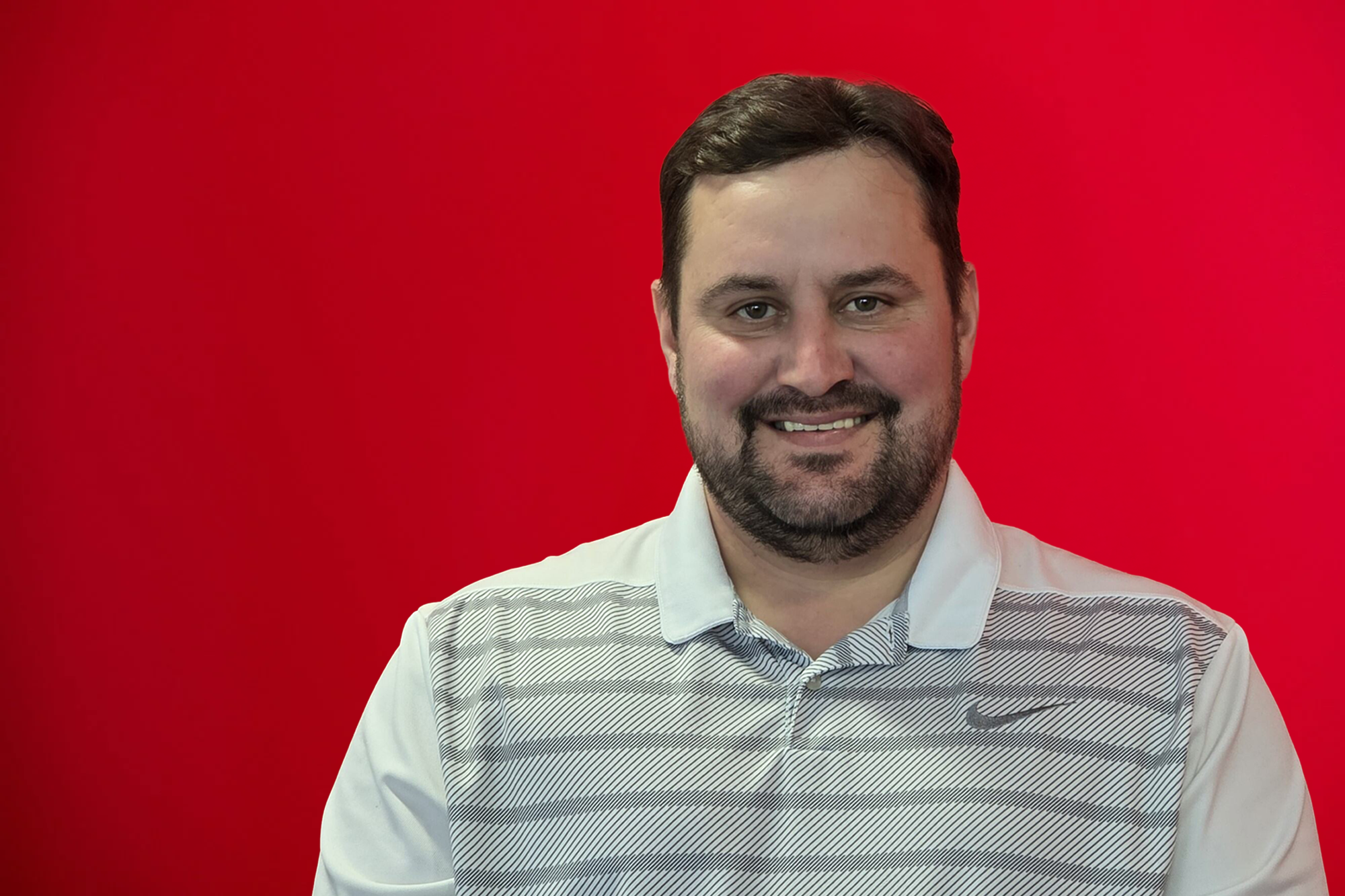 A man with dark hair, a beard, and a mustache, smiling, wearing a white Nike golf shirt, posed against a red gradient background.