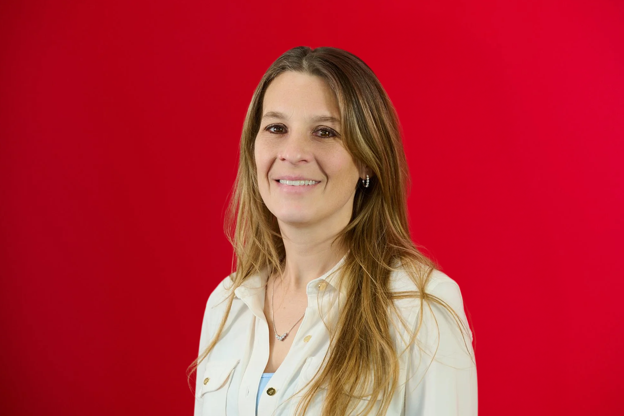 A woman with long light brown hair, wearing a white collared shirt and jewelry, smiling in front of a red background.