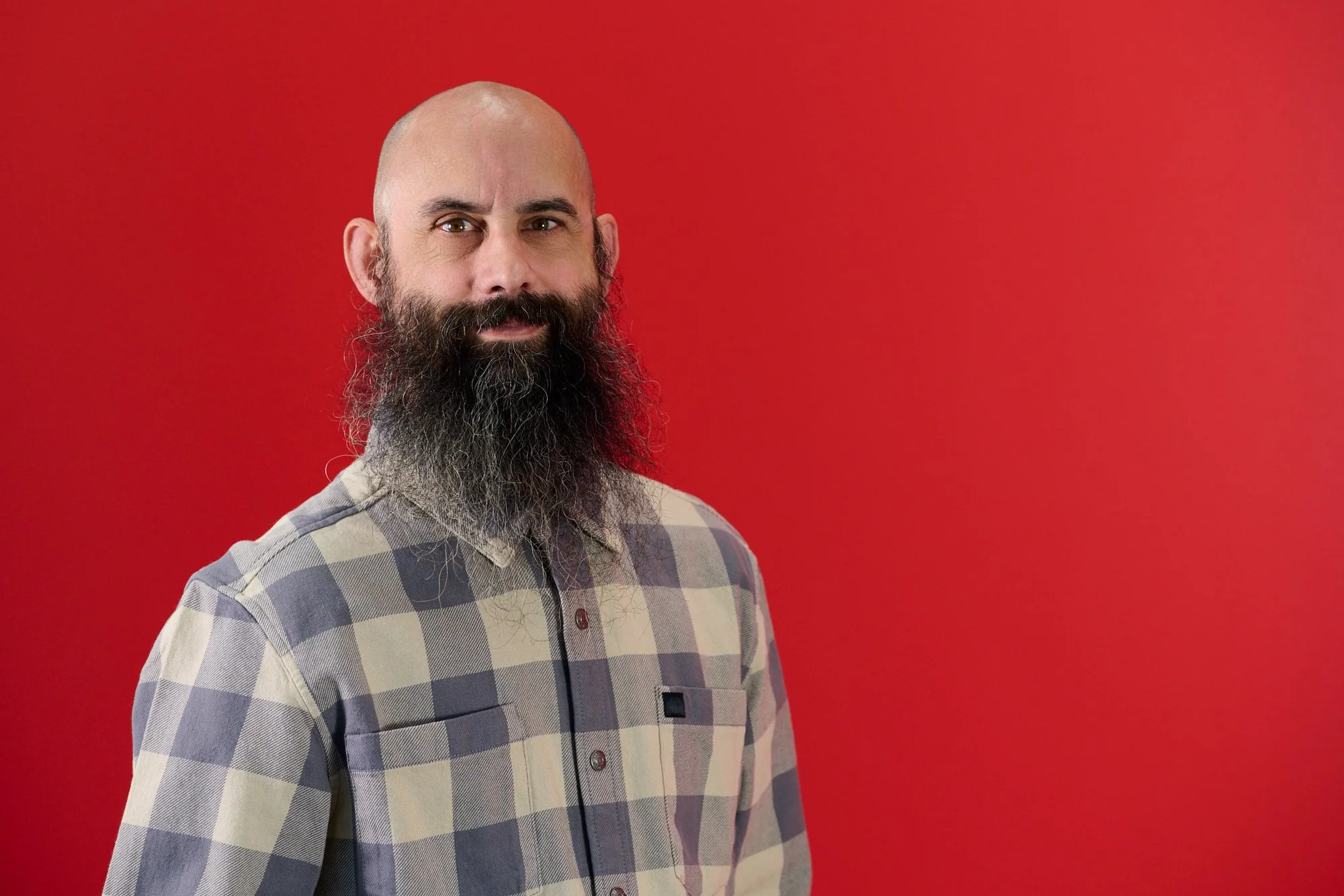 A man with a bald head and a full, long, bushy beard standing in front of a red background, wearing a plaid collared shirt.