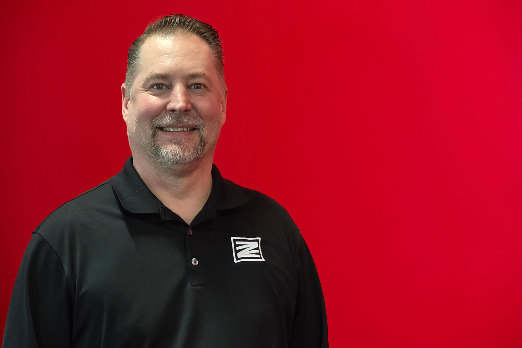 A man with a beard, wearing a gray polo shirt with a logo, smiling in front of a red background.