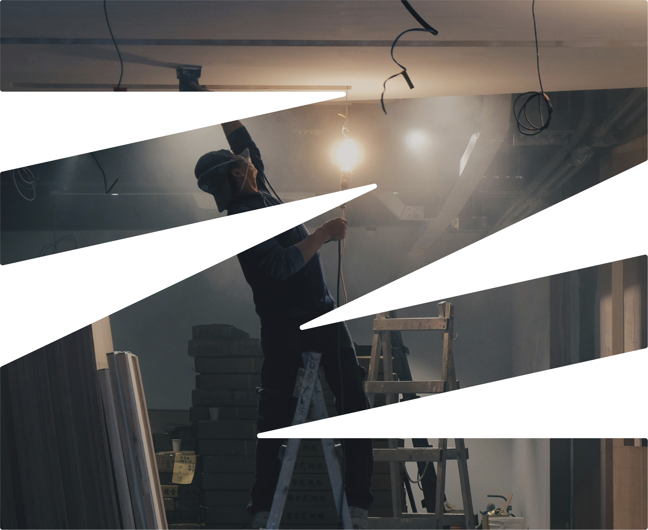 A worker standing on a ladder, installing or repairing electrical wiring on a ceiling with hanging wires and a bright light bulb in an unfinished or under-construction room.