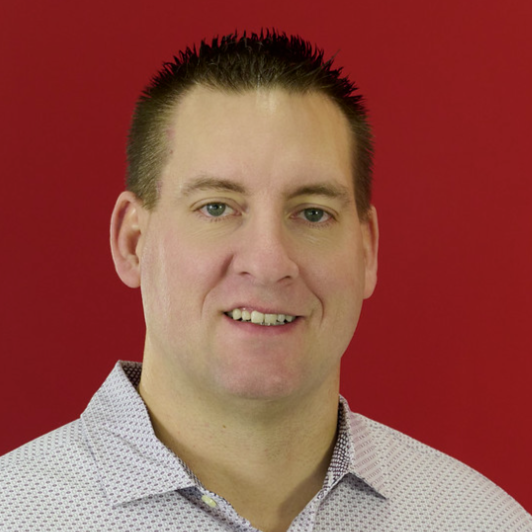 Portrait of a man with short dark spiked hair, wearing a collared shirt with a red background.